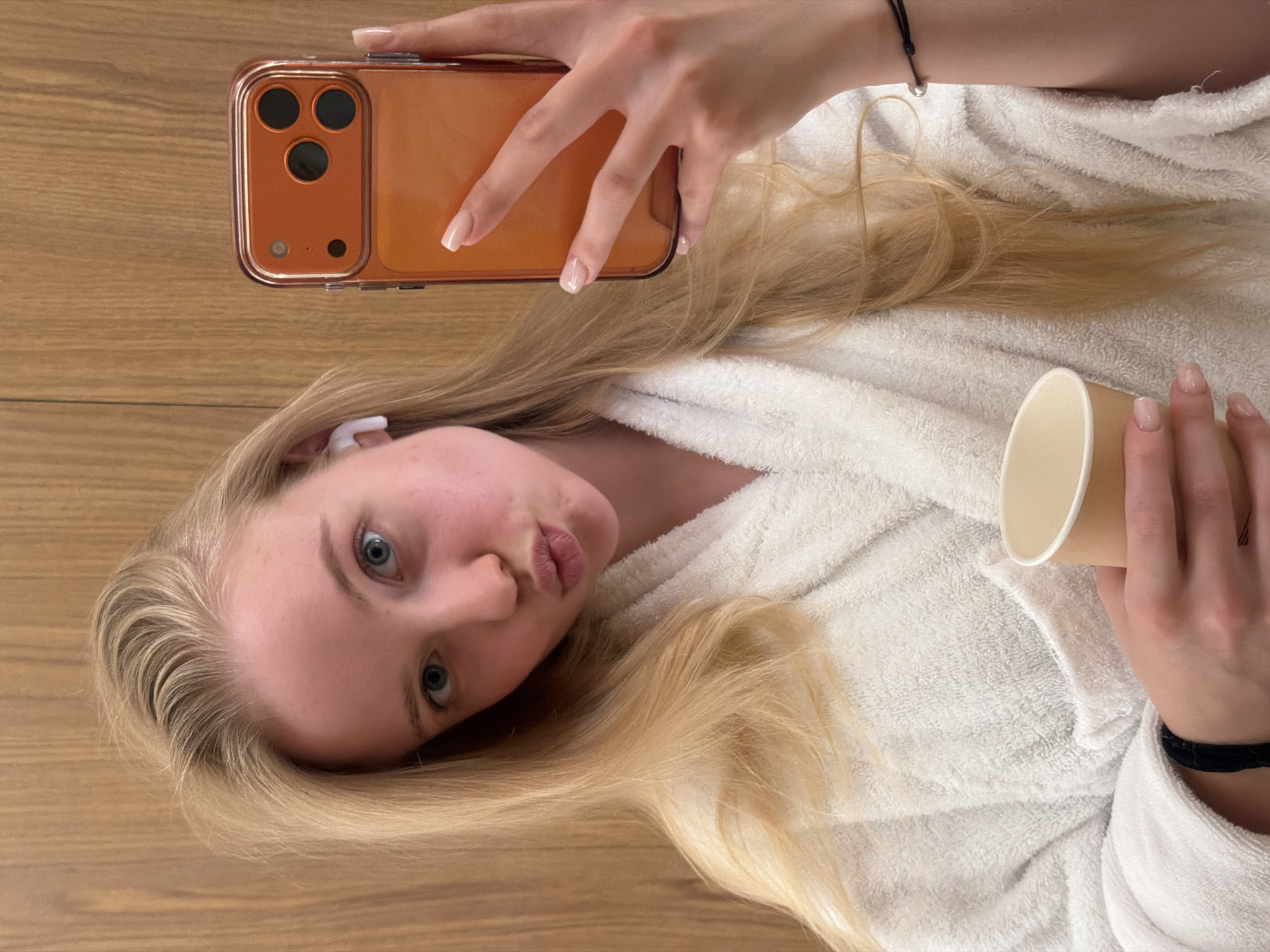 A young woman with blond hair and fair skin is taking a selfie indoors. She is wearing a white bathrobe and an earbud in her left ear. She is holding an orange phone with three camera lenses in her left hand and a beige paper cup in her right hand. She is looking at the camera with her lips pursed. She is lying on a wooden surface. Location is unknown.