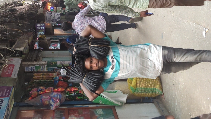 A young man carries two bundles of black, rolled material, secured with light-colored twine, balanced on each shoulder while walking on a paved urban street in Dhaka, Bangladesh. He is in his late teens or early twenties, has dark curly hair, and wears a horizontally striped white and light blue short-sleeved t-shirt with "Never forget" written across the chest, and dark faded jeans. His mouth is slightly open, and he looks straight ahead.
To his immediate left, a shopfront features various brightly colored packaged goods hanging for display, with a large yellow and green woven sack resting on the pavement below. The building behind him and to his left displays a red sign with white Bengali script overhead. In the background, other pedestrians are visible, including a man in a patterned shirt walking away from the camera and another partially visible on the far right holding a blue object. The street is lined with multiple shops and buildings, exhibiting a dense network of black electrical wires crisscrossing overhead. Several signs with Bengali script are visible in the background, including a poster featuring a man's face. The overall scene depicts a bustling street environment.
