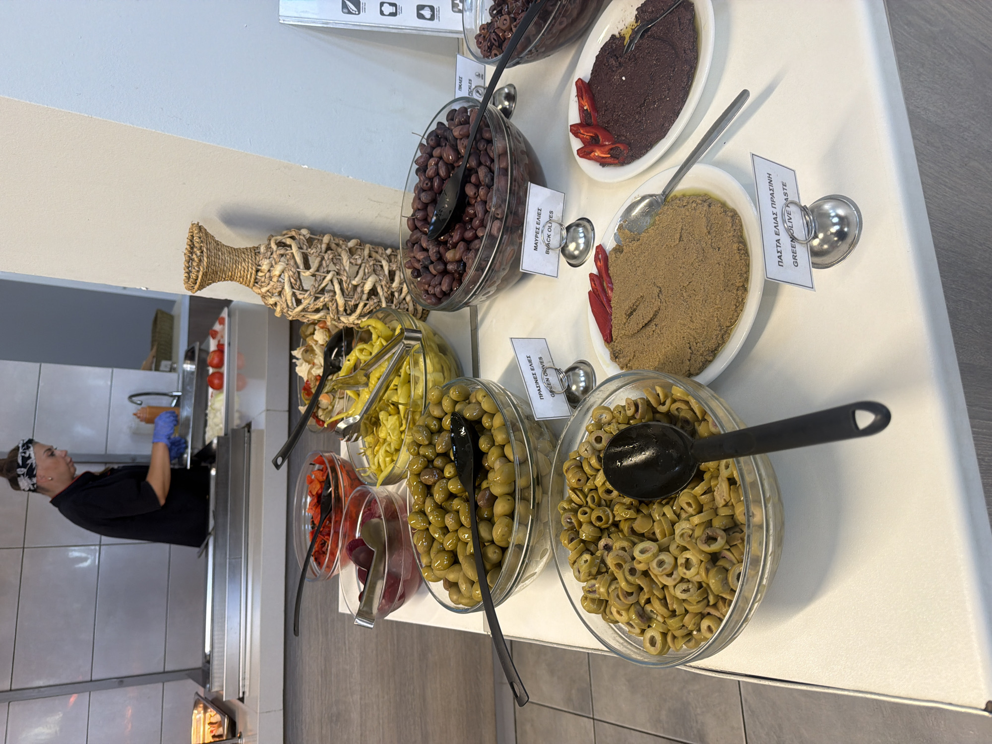 A woman in a black chef's uniform and blue gloves is preparing food at a buffet station in Georgioupoli, Greece. The buffet features various olive dishes in glass bowls, including green olives, sliced green olives, and black olives. There are also bowls of red peppers, yellow pickled peppers, and what appears to be a spread or paste, possibly olive paste, served on white platters. A decorative woven vase is visible on the right side of the buffet. The environment suggests a restaurant or hotel dining area.