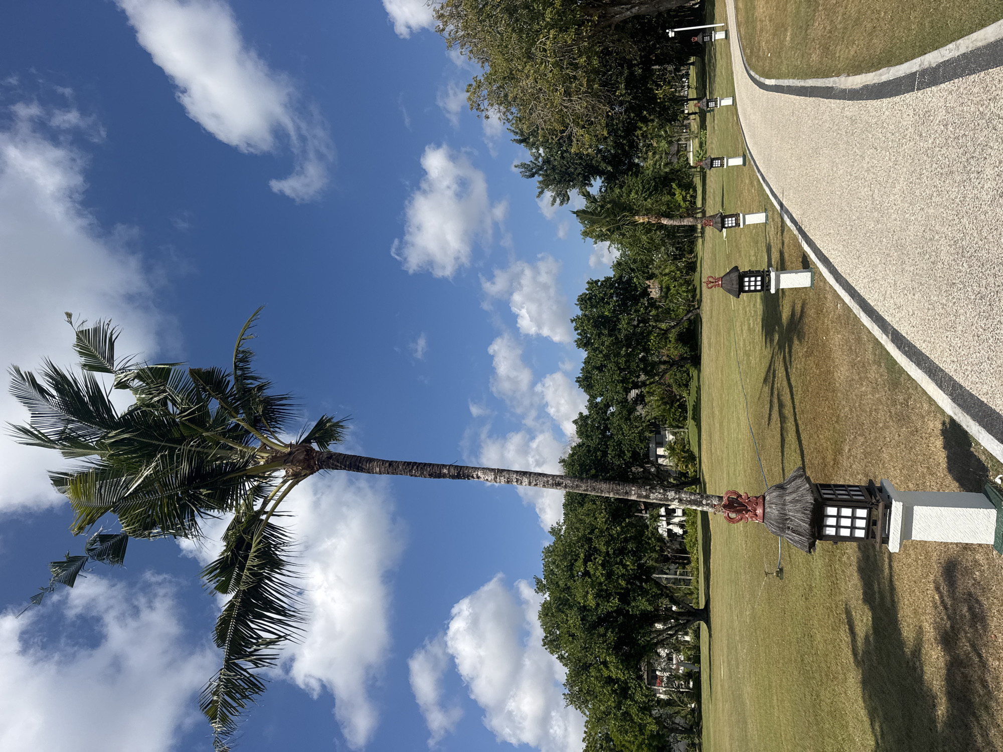 The image depicts a paved pathway curving through a manicured lawn in Kuta Selatan, Indonesia. A tall palm tree stands prominently to the left, its fronds reaching towards a vibrant blue sky dotted with fluffy white clouds. Along the edge of the pathway, a series of decorative lamps, each with a square lantern and a thatched roof topped with a red, crown-like finial, are evenly spaced. The lawn is a lush green, transitioning to a lighter, drier shade in some areas. In the background, a dense line of trees forms a natural backdrop, with hints of buildings visible through the foliage. The overall scene conveys a sense of tropical tranquility and well-maintained landscape, typical of resort areas in Bali.