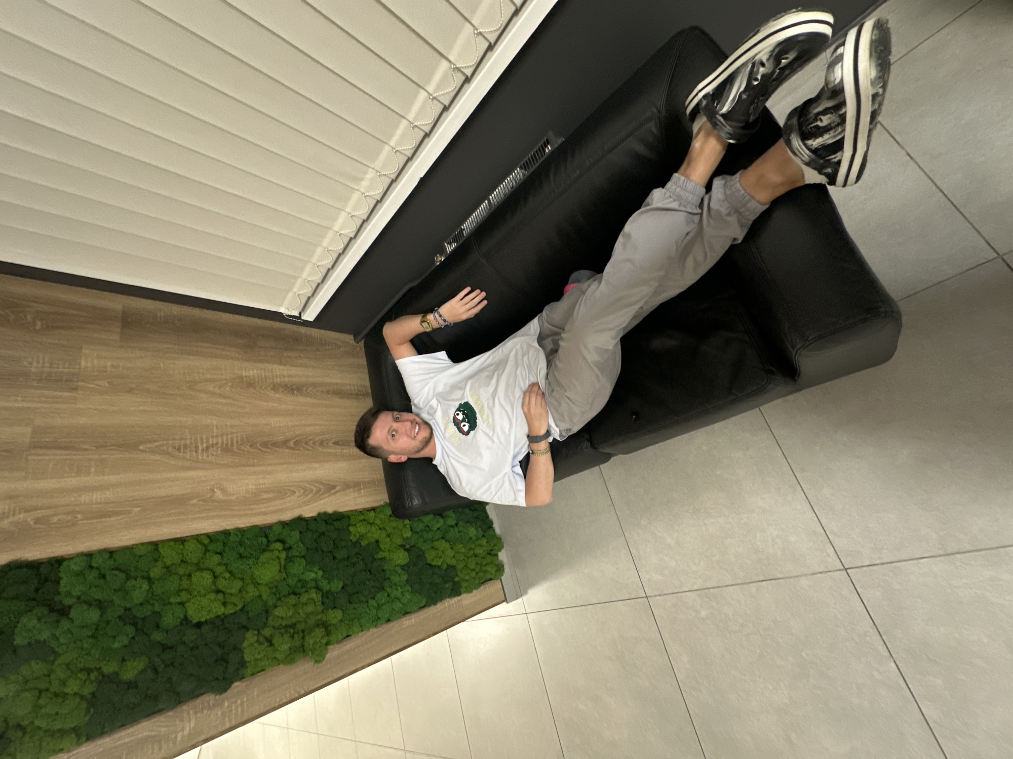 A man is reclining on a black leather sofa. He is wearing a white t-shirt, gray sweatpants, and black-and-white platform sneakers. The sofa is positioned against a wall with white horizontal blinds. Adjacent to the sofa is a wall featuring a strip of green moss. The floor is tiled with gray tiles. The scene appears to be indoors. There is no visible evidence indicating that this scene is specifically in Moscow, Russia.