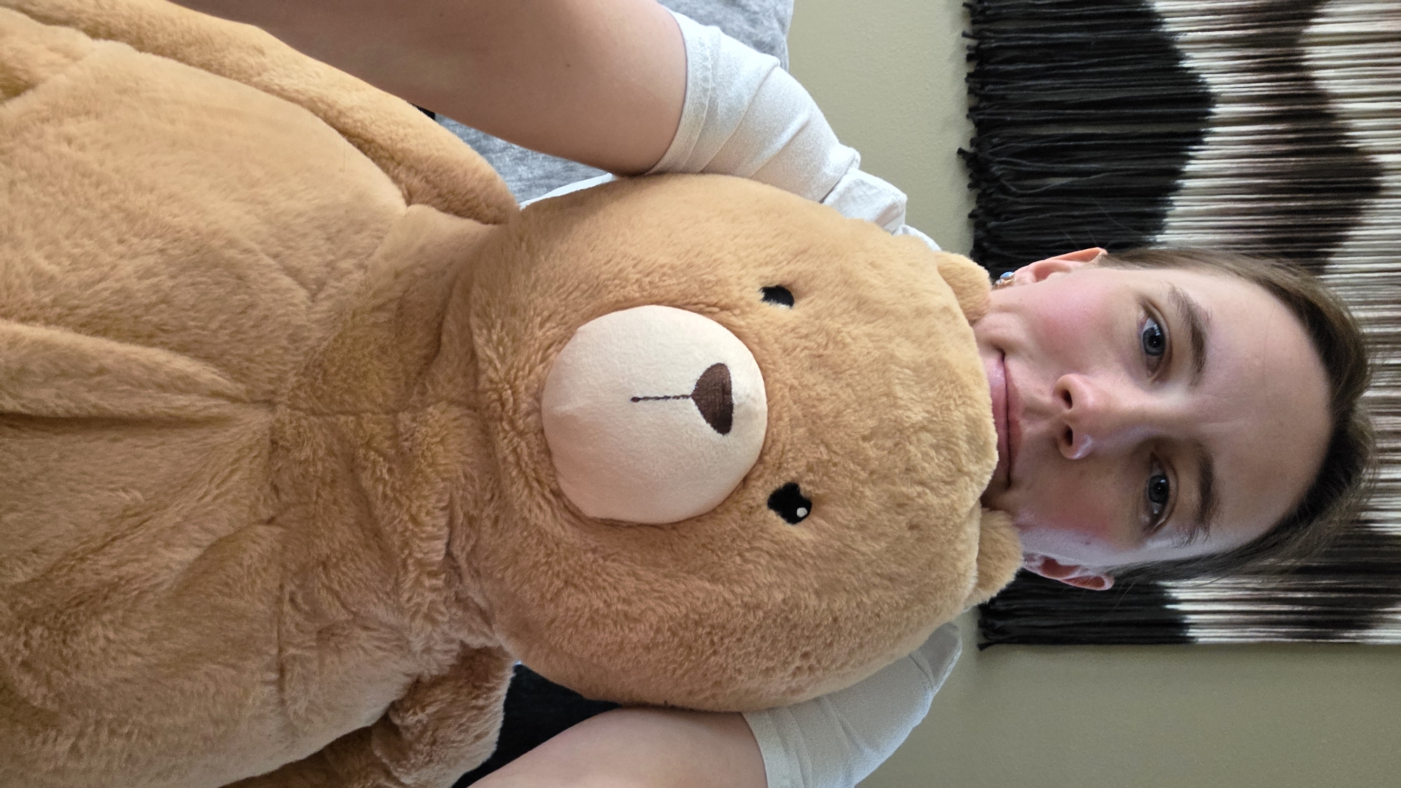 The image features a young woman in a close-up, portrait-style selfie, holding a large, light brown teddy bear. The woman is positioned to the right of the frame, with her head tilted and her gaze directed towards the camera. She has fair skin with rosy cheeks, blue eyes, and light brown hair. She is wearing a light-colored t-shirt. A small, dark earring is visible on her left ear. The teddy bear, which takes up a significant portion of the left side of the frame, has a classic teddy bear design with a lighter-colored snout. Its features, including eyes and a stitched nose, are visible.
The background is divided into two distinct sections. On the left, it appears to be a plain wall or surface, softly lit. To the right of the woman, a textured wall hanging with vertical stripes of black and white (or dark brown and beige) is visible. The lighting suggests an indoor setting, possibly a home or a studio, during daytime. There are no overt indications of the weather.
The image appears to capture a moment of comfort and affection, with the woman embracing the teddy bear. Her expression is gentle and relaxed. The composition emphasizes the connection between the woman and the bear, with the bear's head resting near her face. The setting provides a neutral backdrop that focuses attention on the subjects. No text is visible in the image.