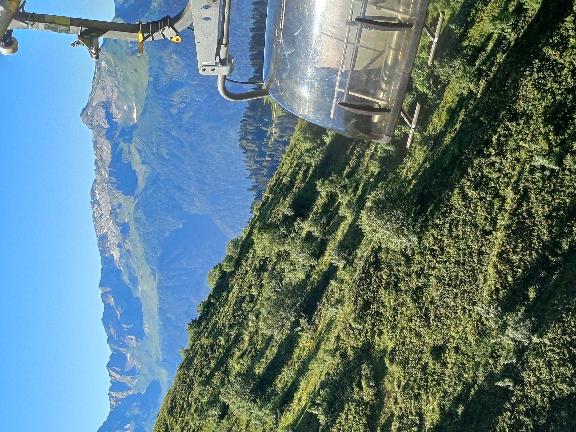 An elevated, wide-angle shot captures a view from a ski lift chair over a lush green mountain slope under a clear blue sky. The ski lift chair, made of metal and transparent plastic, is in the foreground on the right, its mechanical arm visible. The slope, covered in dense green vegetation and scattered trees, descends diagonally from the left to the right. In the background, a range of imposing mountains with rocky peaks, some still holding patches of snow, rises against the horizon. Below the snow-capped mountains, a dense forest of dark green trees creates a contrasting band before meeting the lighter green of the lower mountain slopes. The scene is set in Estosadok, Russia, a mountain resort known for its stunning natural landscapes.