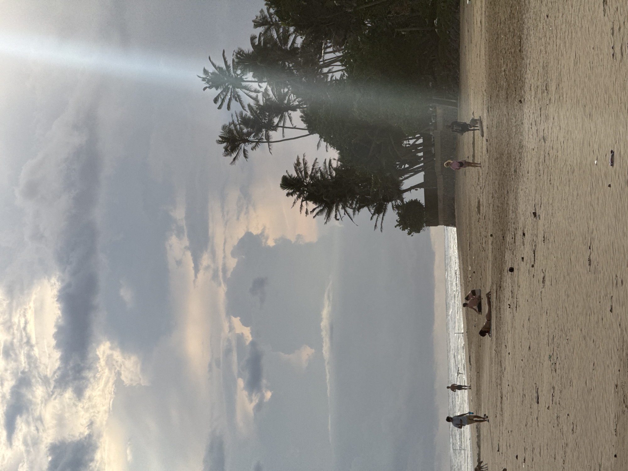 The image captures a beach scene in Kathaluwa, Sri Lanka. The sky is overcast with clouds, and a sunbeam is visible. A line of palm trees is situated between the sky and the beach. Several people are on the sandy beach, some near the water's edge and others further inland. Some people are walking, while others appear to be sunbathing or relaxing. The ocean is visible as a narrow strip.