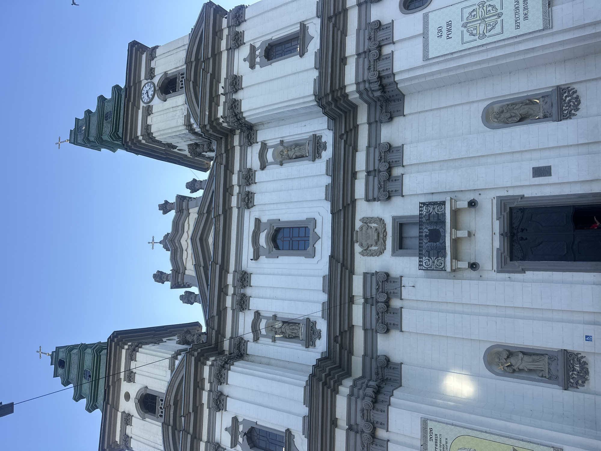 The image shows the Ternopil Theological Seminary, also known as the Church of the Immaculate Conception of the Blessed Virgin Mary. It is a Roman Catholic church and seminary located in Ternopil, Ukraine. The building is a two-towered Baroque structure with a white facade and grey decorative elements. Two large green spires top the towers, each crowned with a cross. A clock is visible on the right tower. Niches on the facade contain statues of saints. Banners are displayed on the left and right sides of the building. The sky is clear and blue. The image is taken from a low angle, looking up at the facade of the building.