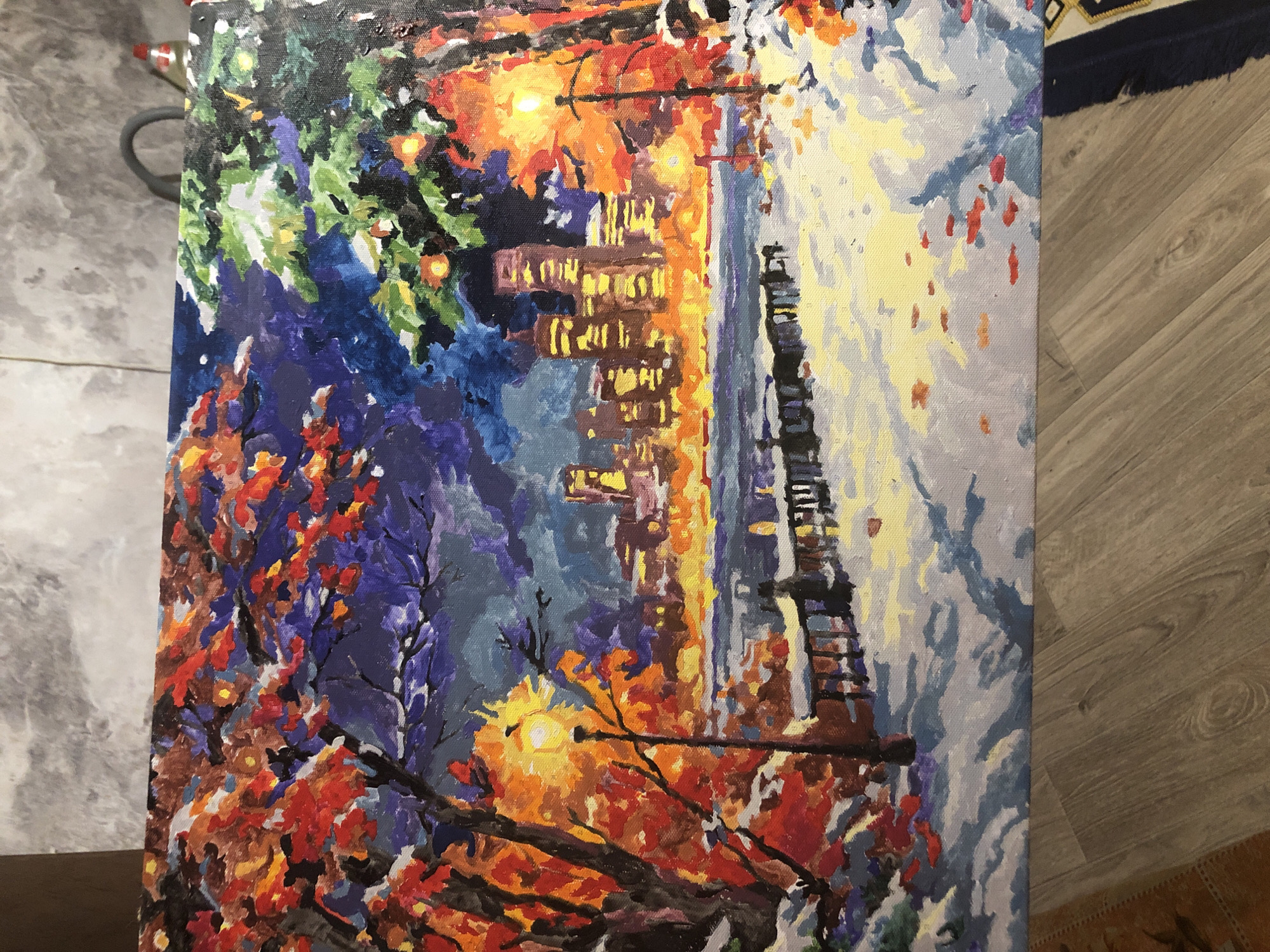 The image is a painting of a winter cityscape at night, depicting a waterfront scene with a city skyline in the background. On the left side of the painting, there are trees with vibrant red and orange leaves, suggesting a late autumn or early winter setting despite the snow. A lamppost illuminates the foreground, casting a warm glow. The middle ground shows a body of water with a railing along the edge, and a snowy path leads towards the water. The background features a cluster of illuminated buildings, forming a city skyline. The sky is dark blue, with hints of purple and clouds. The overall impression is a cold but vibrant night scene. The painting does not depict any specific landmarks or features that would place it in the city of Penza, Russia.