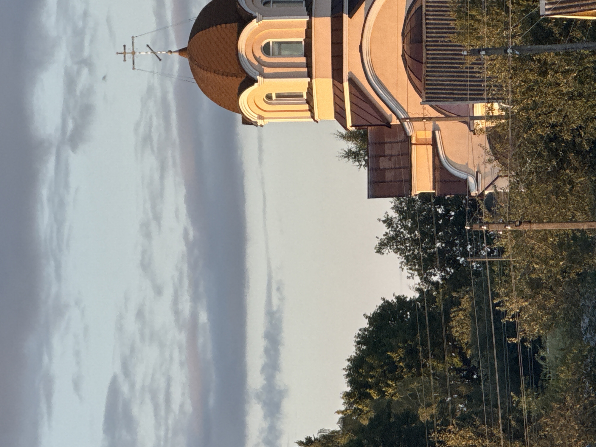 This is a vertical, eye-level outdoor photograph taken in Talachyn, Belarus. The scene features a church with a brown dome and a cross on top, partially visible on the right side of the image. The church is painted in light peach and white colors. The sky above is a pale blue with scattered clouds. In the background, there are trees and power lines. The lighting suggests it might be sunset or sunrise, as the building is illuminated with a warm, golden light.