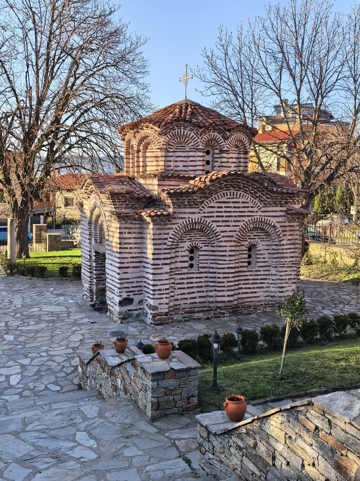 The image depicts a small, brick church with a red-tiled roof and a cross on top. The church is surrounded by trees and greenery. The building is made of red and white bricks, arranged in horizontal layers. The roof is made of red tiles. A stone path leads to the church. The location is in the city of Unknown, Unknown.