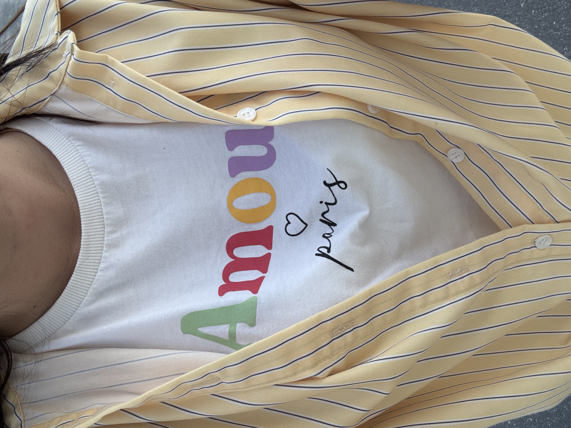 A close-up, eye-level shot captures the upper torso of an individual wearing a white t-shirt layered under an open, yellow and black striped button-up shirt. The t-shirt features the word "Amour" in colorful, varied fonts: "A" in green, "m" in red, "o" in orange, "u" in purple, and "r" in a lighter purple/pink. Below "Amour," a small black outlined heart is positioned, followed by the word "paris" in black, flowing script lowercase letters. The t-shirt has a ribbed crew neckline. The outer shirt is characterized by thin, vertical black stripes on a pale yellow fabric, with several white buttons visible along the front placket, indicating it is worn unbuttoned. Dark hair is visible on both sides of the subject's neck and shoulders. A portion of the subject's skin is visible above the t-shirt's neckline. The background is an out-of-focus, uniform light gray surface, providing no specific environmental details.