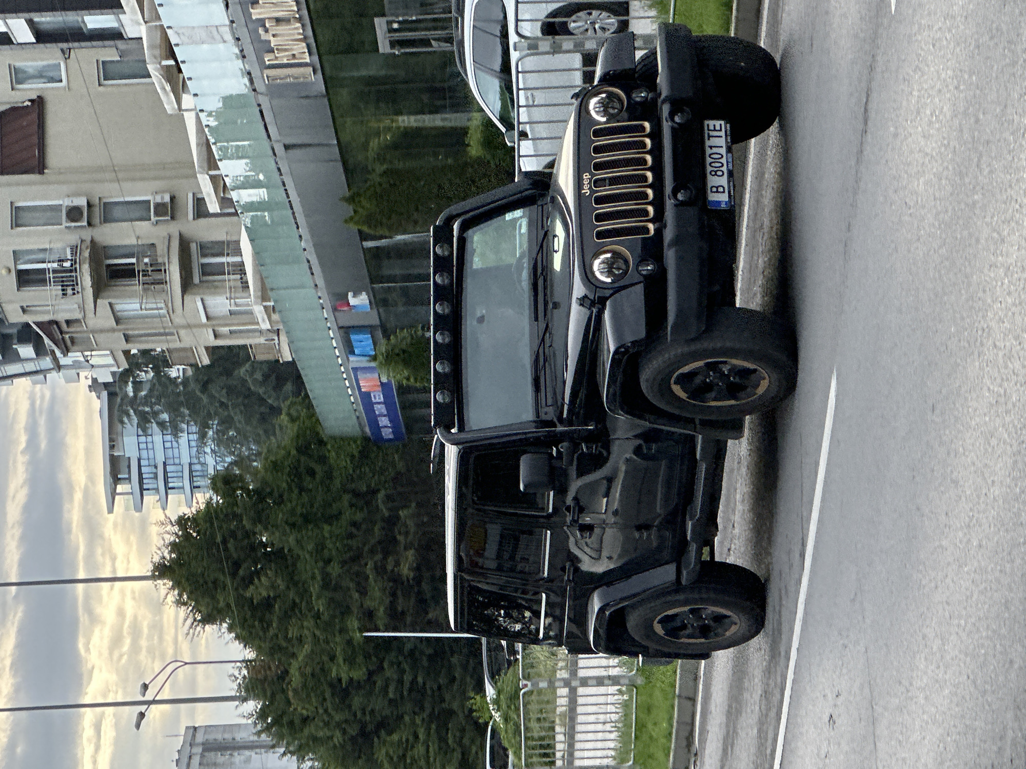 The image shows a black Jeep Wrangler parked on the side of a road in an urban setting. The license plate reads "B 8001 TE." Buildings are visible in the background, suggesting the car is in a city. Based on the license plate and architectural styles, the location is likely Varna, Bulgaria. No people are visible in the frame. The car is the primary subject, parked on a paved road with a white dividing line. A grassy area with a fence is visible on the edge of the road. The sky is overcast.
