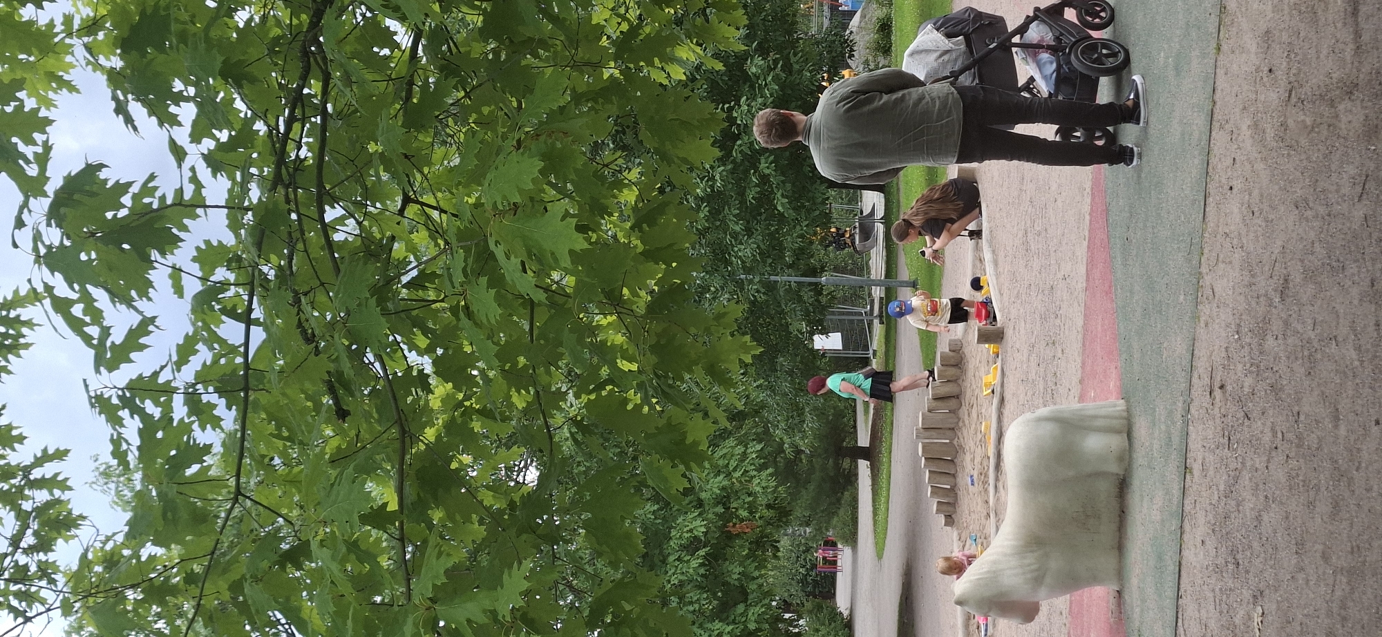 The image shows a playground in Lahti, Finland, under a canopy of green leaves from large trees. In the foreground, a white, stylized sculpture of a horse is visible. To the right, a man stands looking away from the camera, next to a stroller with a person sitting in it. In the midground, a woman in a green shirt and black skirt stands on a wooden obstacle. Two young children are playing in a sandpit; one is on their knees, and another is standing. Further in the background, there are more trees and playground equipment, including a yellow excavator. The ground is covered in sand and a reddish-green surface. The sky is overcast.