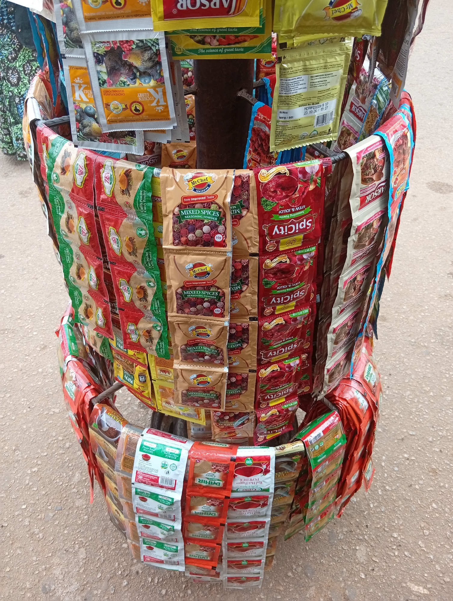This media file shows a close-up, eye-level view of a colorful display of seasoning packets mounted on a metal stand. The packets are arranged in multiple tiers, with some hanging vertically and others displayed horizontally. The stand appears to be placed outdoors, possibly at a market stall, given the rough, sandy ground visible in the background. The lighting suggests it is daytime, with no strong shadows indicating a specific time.
The majority of the visible items are small, individually wrapped seasoning packets, predominantly in red, green, yellow, and brown packaging. Several brands are identifiable, including "Mr.Chef," "SpiCity," "ARSOR," and "DAHIR." The packets feature various illustrations and text, with prominent labeling for "MIXED SPICES," "STEW & JOLLOF," "Curry Powder," and "CHICKEN FLAVOR SEASONING POWDER." Some packets have explicit text such as "New Improved Taste," "The science of great taste," and ingredient lists and nutritional information. The overall impression is one of a vibrant, commercial display of food spices. No people are directly visible in the frame, but the context implies a vendor-customer interaction is likely. The scene is bustling with the colorful variety of the product packaging.