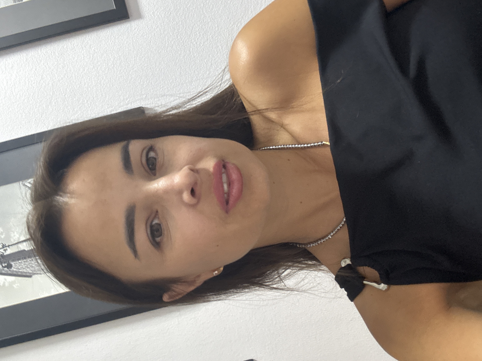 A medium-shot, indoor, chest-up, front-facing portrait shows a woman with medium brown hair and brown eyes. She has light-colored skin and is wearing pink lipstick. She is wearing a black off-the-shoulder top with silver accents on the straps and a silver necklace. She is indoors, with a white textured wall behind her and a framed picture on the wall to the left.