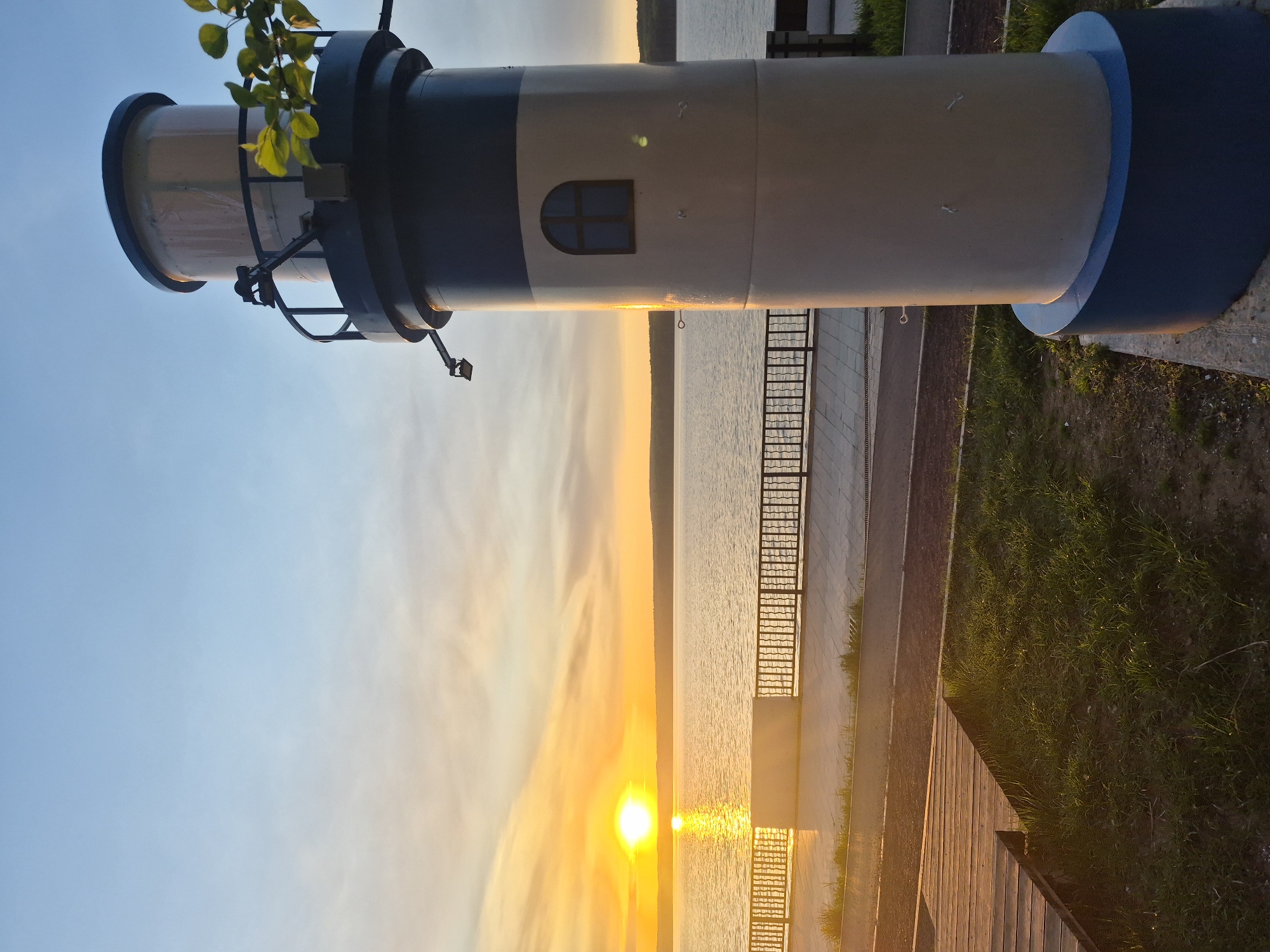 The image captures a lighthouse replica, an observation tower or a viewpoint in the city of Chaikovsky, Russia, likely situated on the Kama River embankment. The lighthouse structure is positioned horizontally in the frame, with the top portion showing a light yellow hue and the lower portion showing a dark blue color. A window is visible on the side. In the background, the setting sun is reflected in the water, creating a brilliant golden light. A walkway borders the water, and there is a metal railing in the scene. Vegetation (grass and green leaves) is visible around the structure.