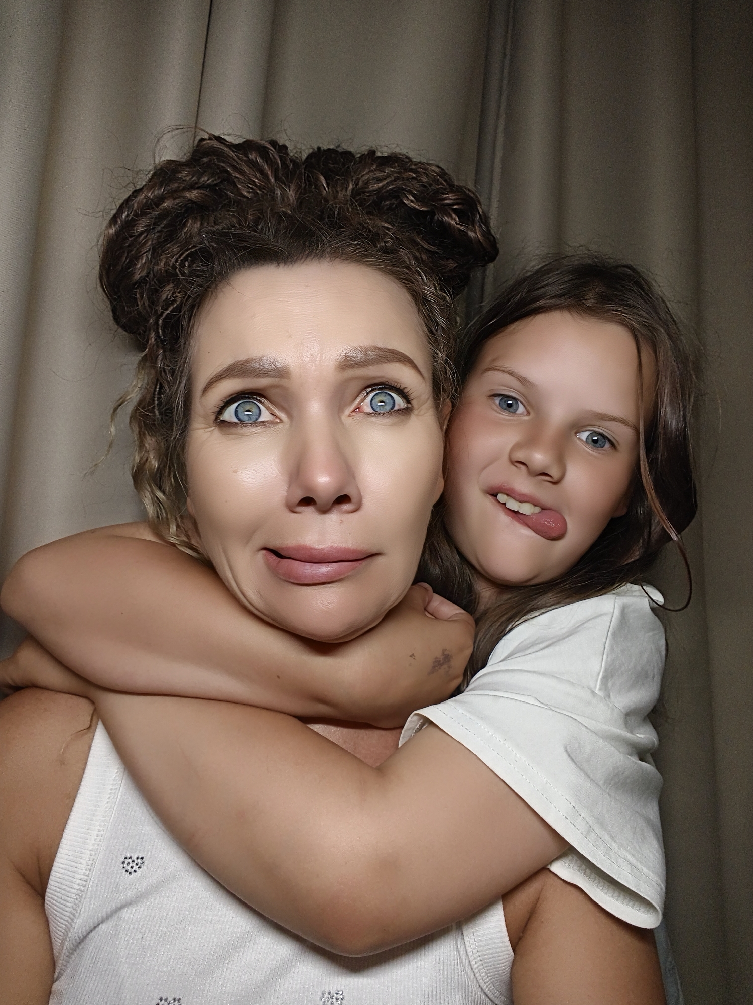 An extreme close-up shot shows a woman and a girl embracing, with the girl's arms wrapped around the woman's neck from behind. The woman is looking up and to the right with wide, surprised eyes, her mouth slightly open. Her hair is pulled up in two messy buns. The girl is sticking her tongue out and looking directly at the camera. She is wearing a white t-shirt. The woman is wearing a white tank top with a subtle pattern. The background is a muted, textured curtain. The lighting is soft, creating a slightly diffused effect.