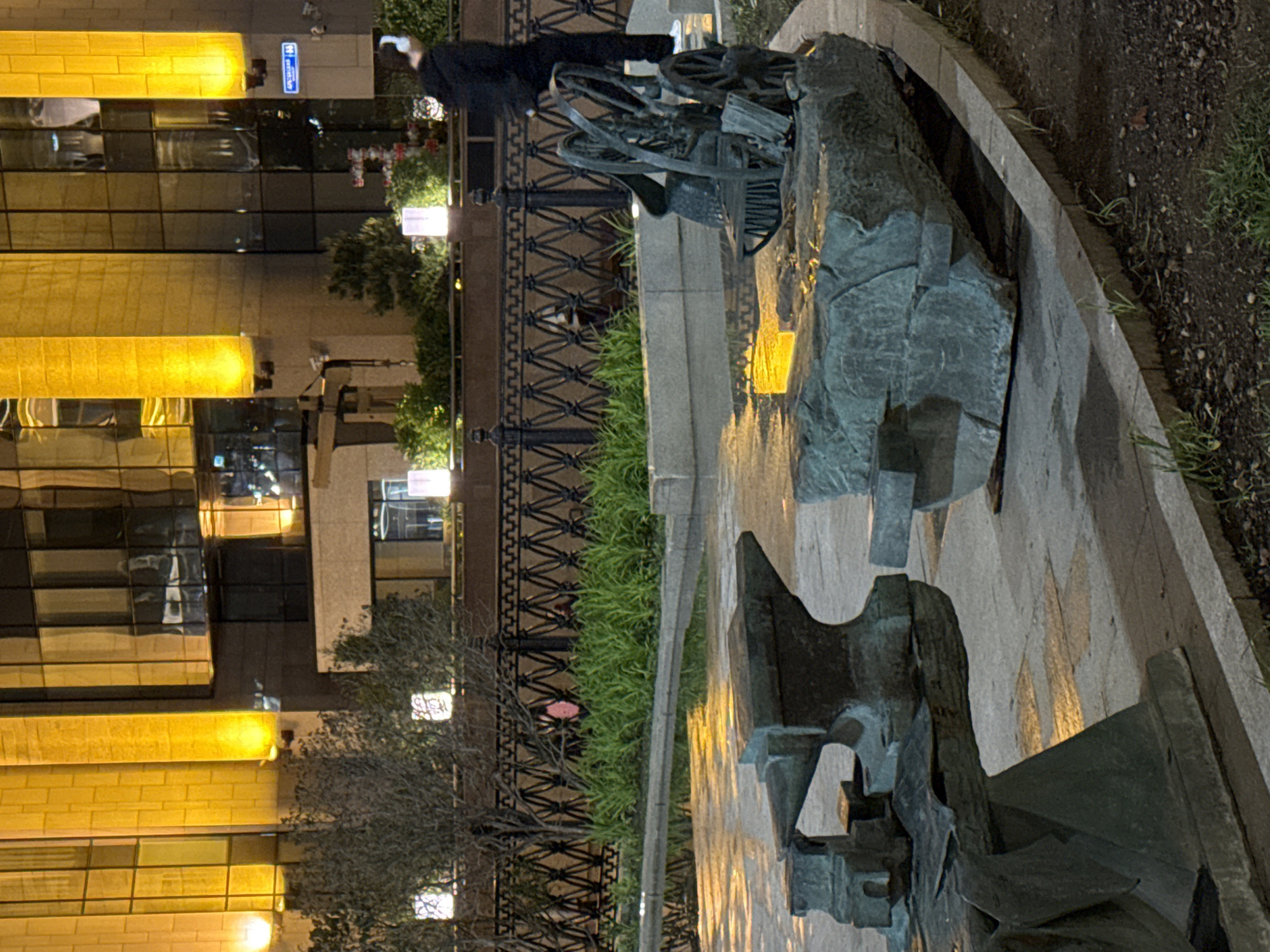 A nighttime, ground-level shot depicts a cityscape and a monument, rotated 90 degrees clockwise. A person stands behind a black metal fence near the monument, which is constructed of rough, dark gray stone and metal. The cityscape, featuring buildings with illuminated windows, is in the background. The foreground consists of stone paving and grass. A building sign is visible. The location is Moscow, Russia.