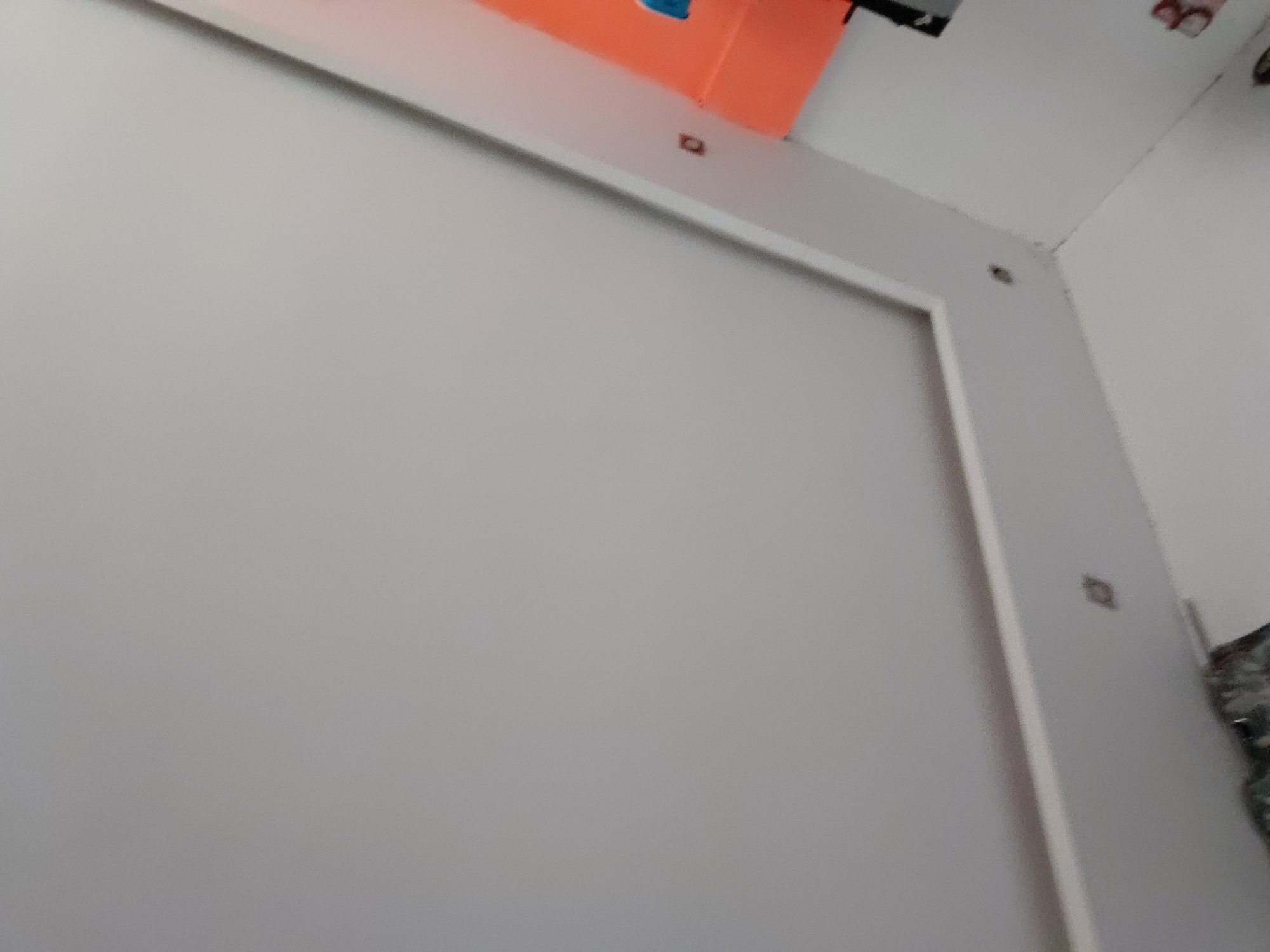 The image captures an indoor scene, primarily featuring a large, light gray or off-white rectangular panel dominating the foreground. This panel is outlined by a white frame or border, suggesting it might be a screen, a whiteboard, or a wall-mounted fixture.
In the upper background, an orange, box-like object is visible, positioned on a white surface or shelf. To its left, a dark, indeterminate object is partially seen. The walls and ceiling of the room are also light gray or off-white, with a corner visible in the upper right.
Several small, dark, rectangular or square-shaped elements are present on the white frame and the upper wall, possibly indicating screw heads, mounting points, or small decorative details. On the far right edge of the image, a dark, textured object, possibly fabric or a bag, is partially visible.
No specific activity is taking place. The scene appears static, capturing a section of an interior space. The lighting is even, suggesting either diffused natural light or artificial illumination, without indicating a specific time of day. No text is visible in the image. The composition is slightly angled, giving a dynamic feel to the otherwise static subjects.