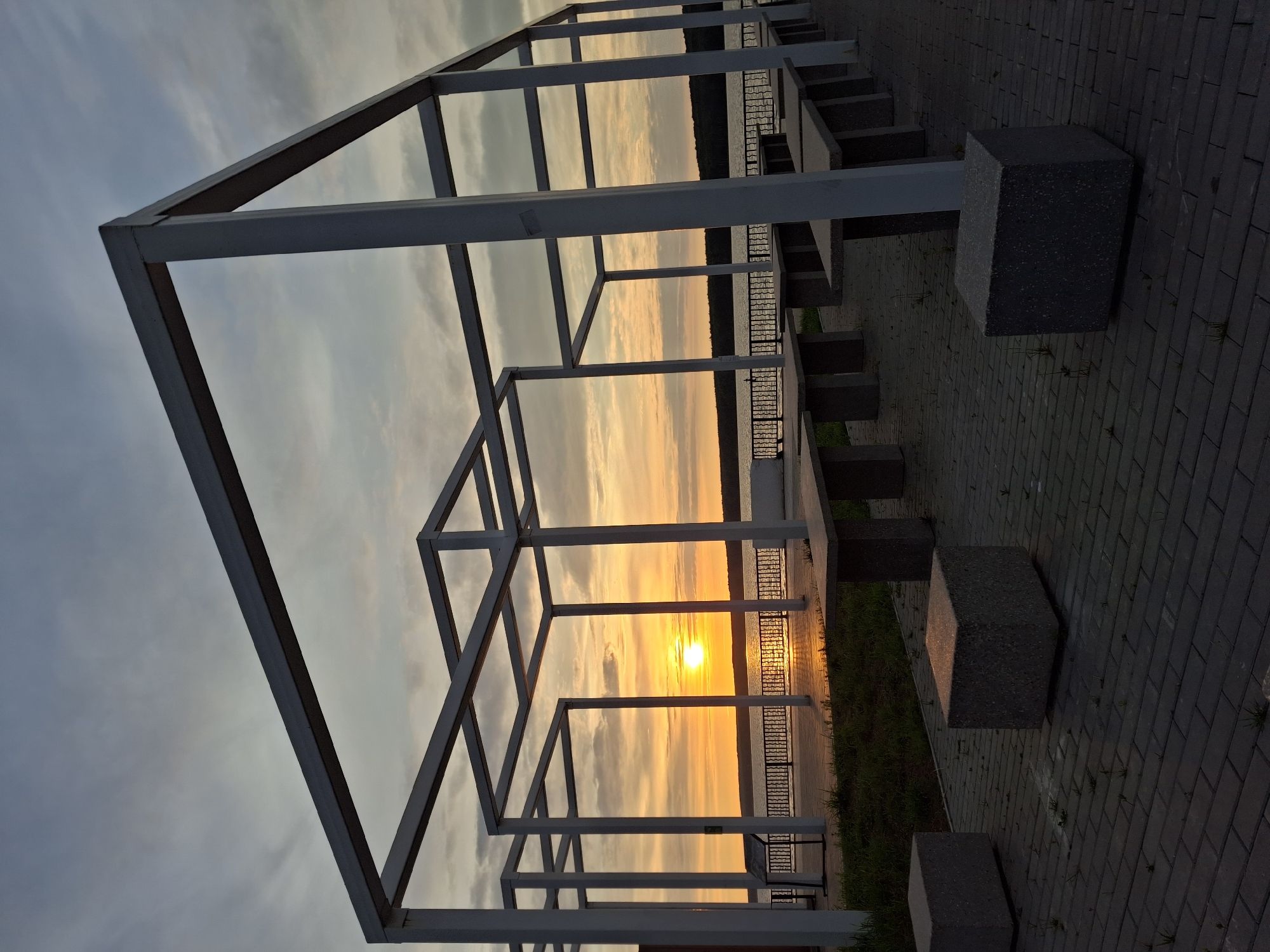This media file depicts a sunset scene in Chaikovsky, Russia. The primary subjects are an abstract, open-air metal structure with a geometric design, and the natural setting of a waterfront. The structure, made of white metal beams forming cubic shapes, is positioned in the foreground, framing the view of the sunset. The sunset is a prominent element, with the sun a bright orb on the horizon, casting a warm orange and yellow glow across the sky and reflecting on the water.
The scene is set on a paved walkway, likely a promenade or embankment, indicated by the dark gray brickwork and the presence of concrete blocks that appear to be seating or design elements. A metal railing separates the walkway from the water. Beyond the railing, a body of water is visible, with a treeline along the far bank.
The time of day is clearly sunset, as evidenced by the low position of the sun and the vibrant colors in the sky. The weather appears to be clear with some wispy clouds. The overall emotion evoked by the image is one of tranquility and natural beauty. There is no visible text in the image.