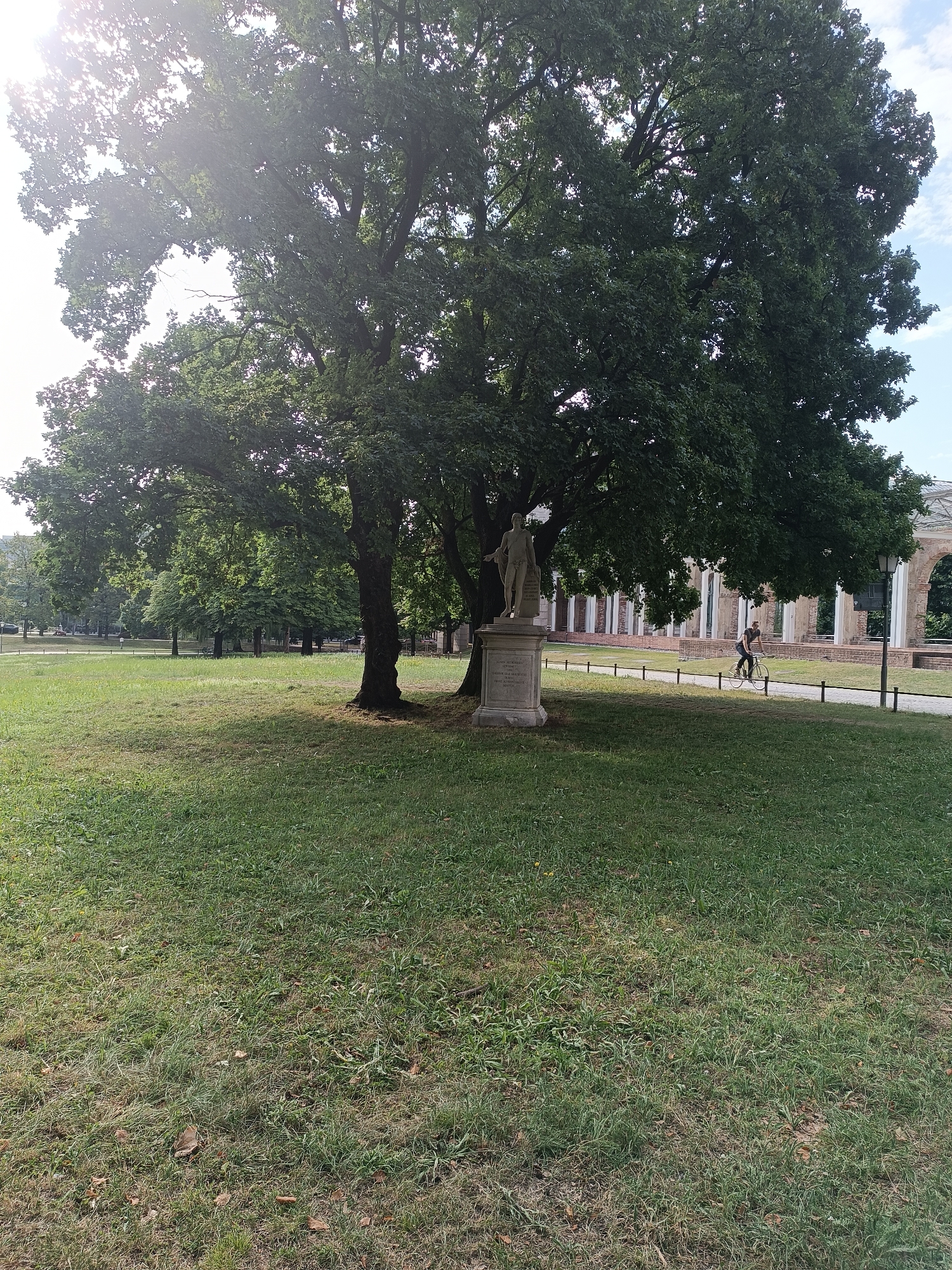 The image depicts a park scene in Munich, Germany. In the foreground, a large, mature tree with dense green foliage dominates the left side of the frame, with its trunk partially visible. Sunlight filters through the leaves, creating a bright, somewhat washed-out effect in the upper left corner. In front of the tree, on a stone pedestal, stands a classical statue of a male figure.
A paved pathway runs horizontally across the middle ground, bordered by a low black fence. To the right of the pathway, a person is riding a bicycle. Behind the pathway, there is a long, colonnaded structure with white pillars, appearing to be part of an old building or ruins. The area around the pathway and the structure is grassy, with some areas appearing drier than others. In the distance, more trees and some buildings are visible, suggesting an urban park setting. The sky is partly cloudy.