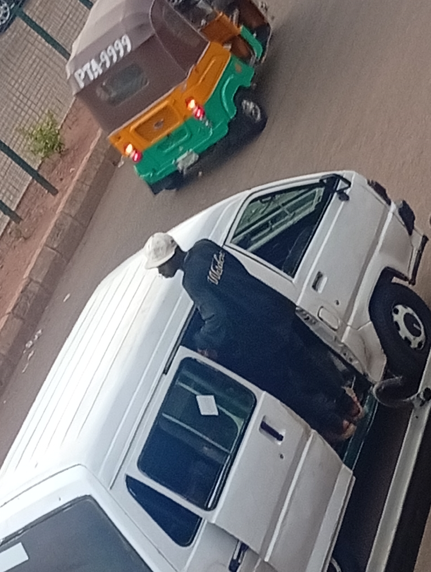 A man is standing next to a white van with its door open, parked on a paved surface. In the background is a green, yellow, and brown auto rickshaw. The rickshaw's license plate reads "PTA-9999." The location appears to be in a city, but the exact location and city name are unknown.