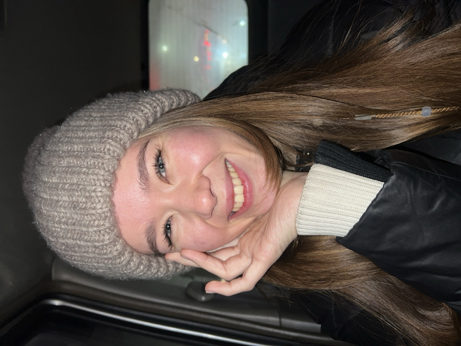 A young woman smiles broadly, wearing a beige knit hat and a black jacket with a cream-colored sweater cuff visible. Her long brown hair cascades over her shoulder. She is partially obscured by what appears to be a car window. Her hand is near her face. Light reflects off her skin and the knit hat. The background is dark with some blurry lights.