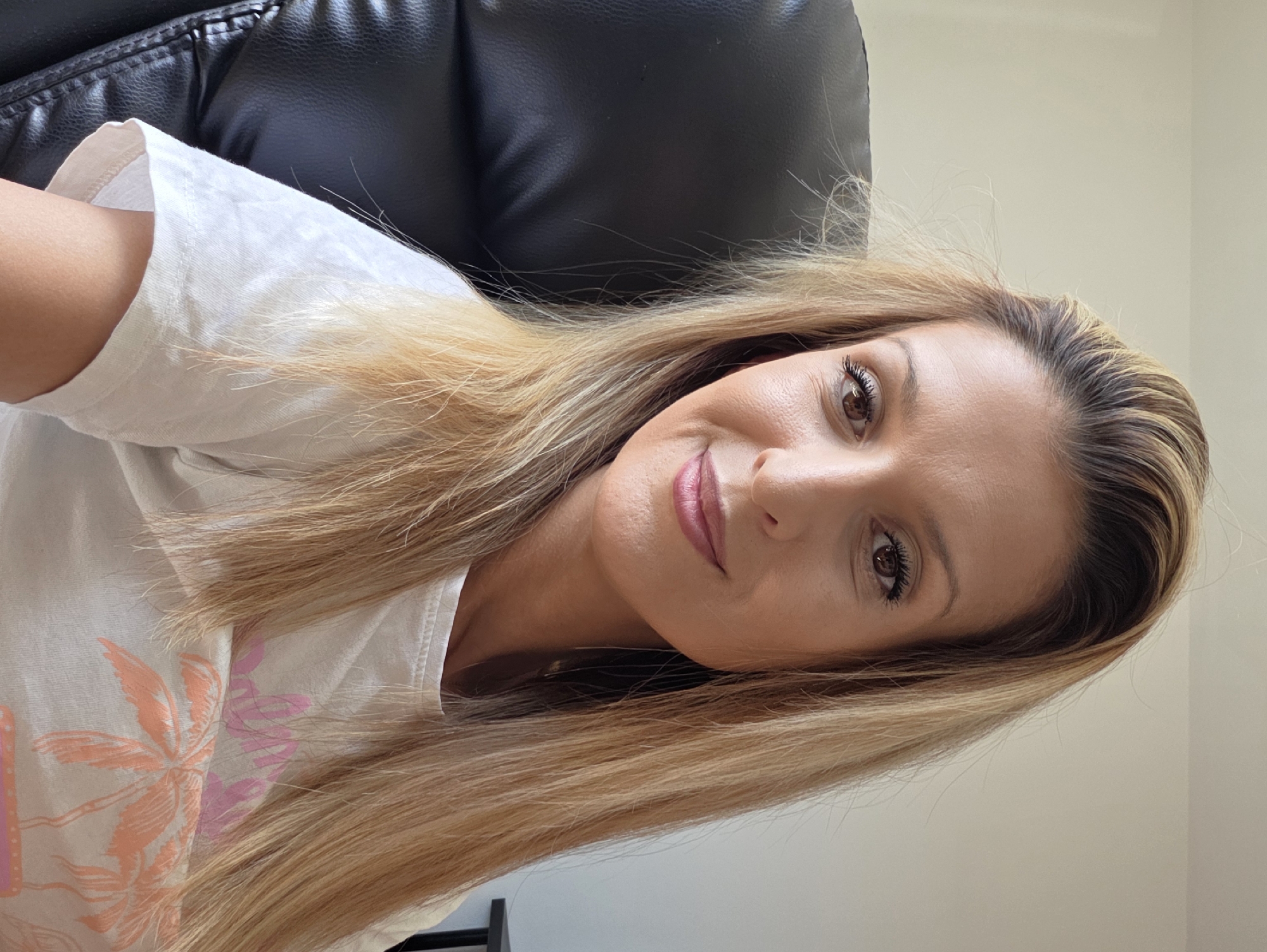 A close-up, head-and-shoulders shot shows a blonde woman with brown eyes, smiling and looking at the camera. She is wearing a white t-shirt with a palm tree graphic. She is seated in a black leather chair, indoors, with a neutral-colored wall behind her. The image does not provide any information about the city of Munich, Germany.