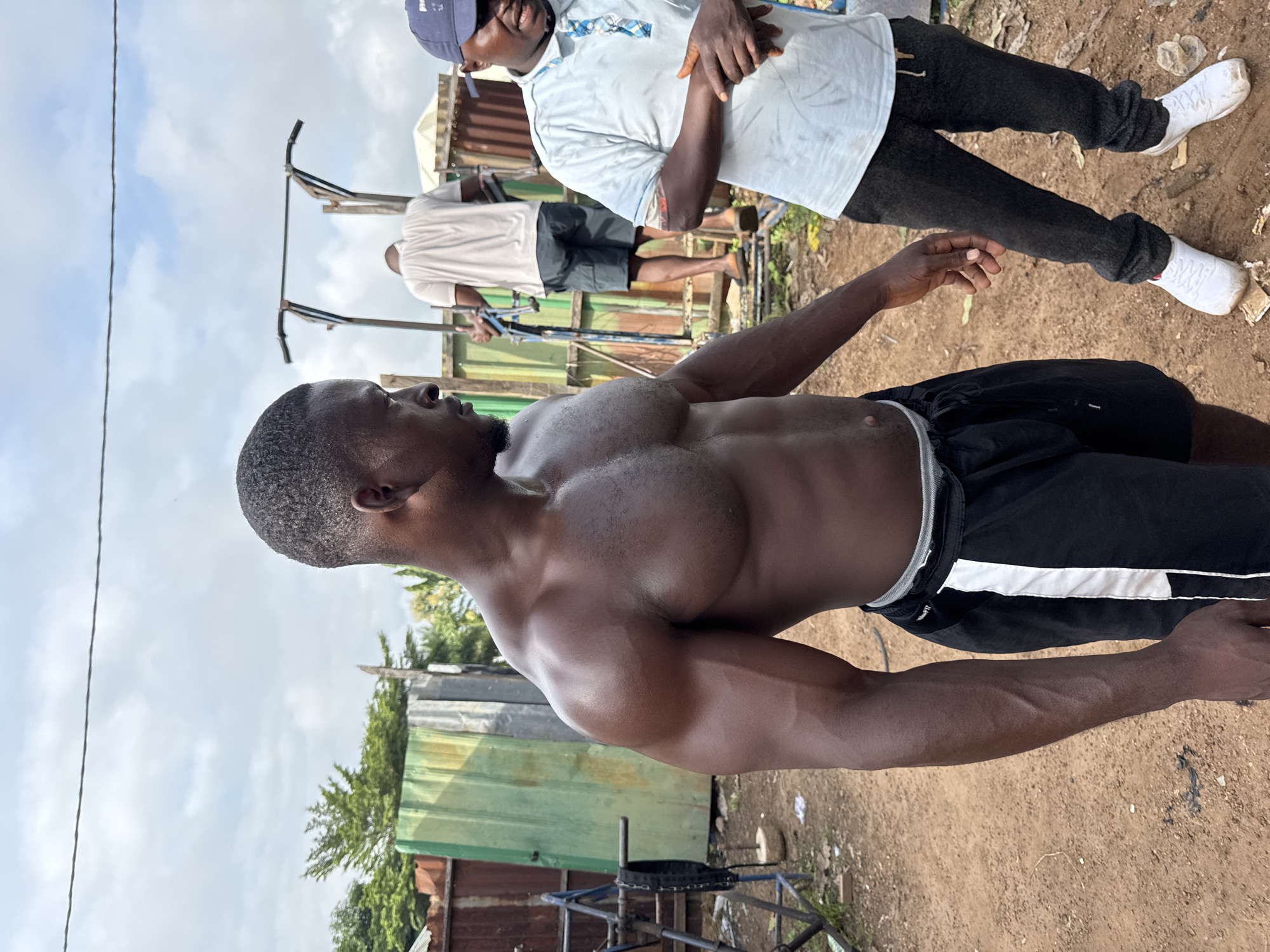 In Akwanga, Nigeria, a muscular man is photographed from the chest up, looking to his right. He is shirtless, showcasing his well-defined physique, and is wearing black athletic shorts with white stripes. To his right, another man is visible from the waist down, wearing a light blue collared shirt, dark pants, and white sneakers. He appears to be speaking. In the background, a pull-up bar structure stands, with another man in a white t-shirt and shorts using it. The environment is outdoors, with corrugated metal structures and foliage visible, suggesting a rural or semi-urban setting. The ground is dirt and gravel. The sky is overcast.