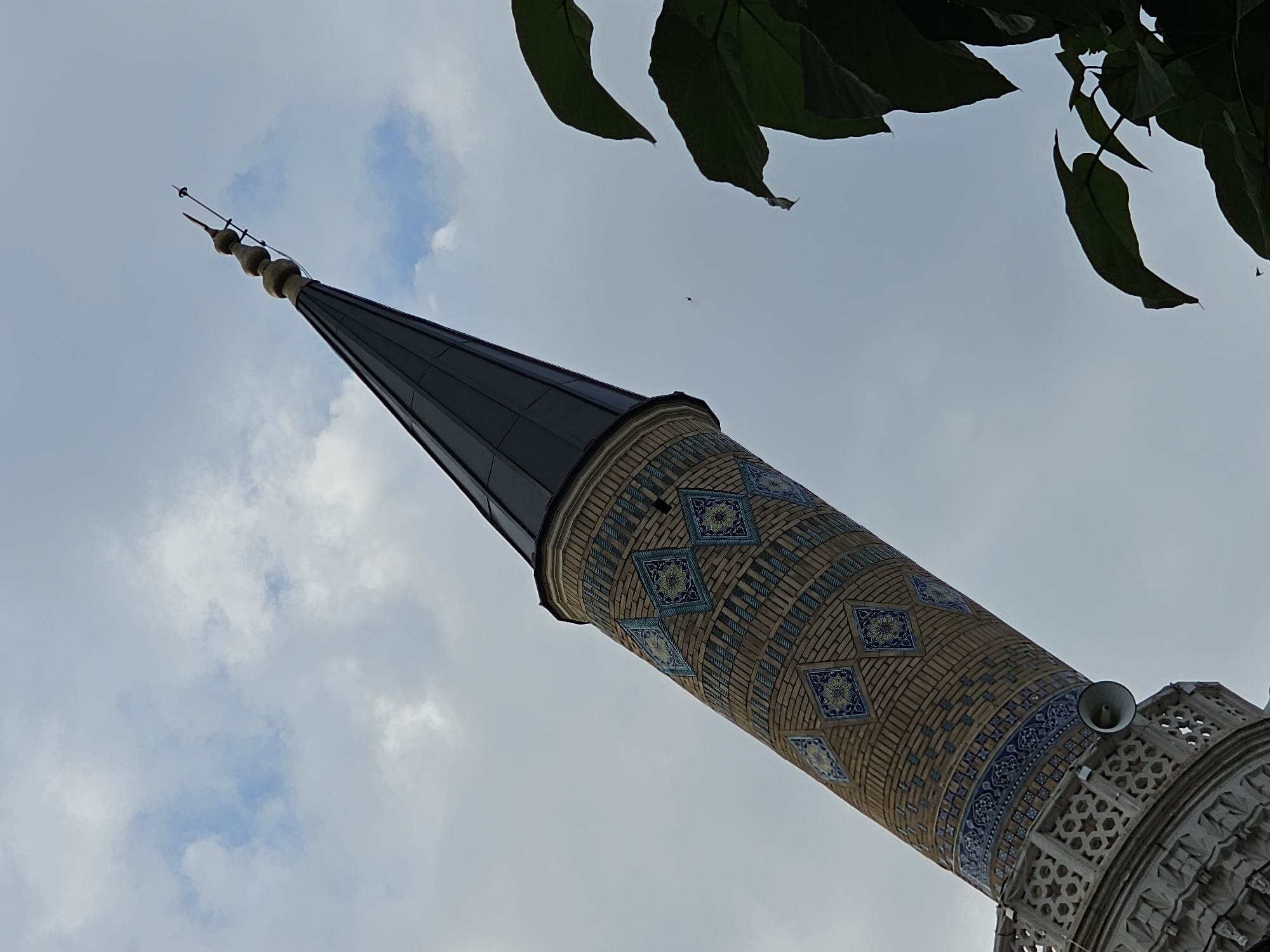This is a low-angle, upward shot of a minaret in the city of Özgön, Kyrgyzstan. The minaret, constructed of brick and adorned with intricate blue and yellow tilework, tapers upwards to a dark conical roof topped with a finial. A balcony with a decorative railing encircles the lower part of the minaret, and a speaker is mounted on the railing. The sky is overcast with scattered clouds, and the upper right portion of the image is partially obscured by the dark green leaves of a tree. The overall impression is one of architectural grandeur against a muted sky.