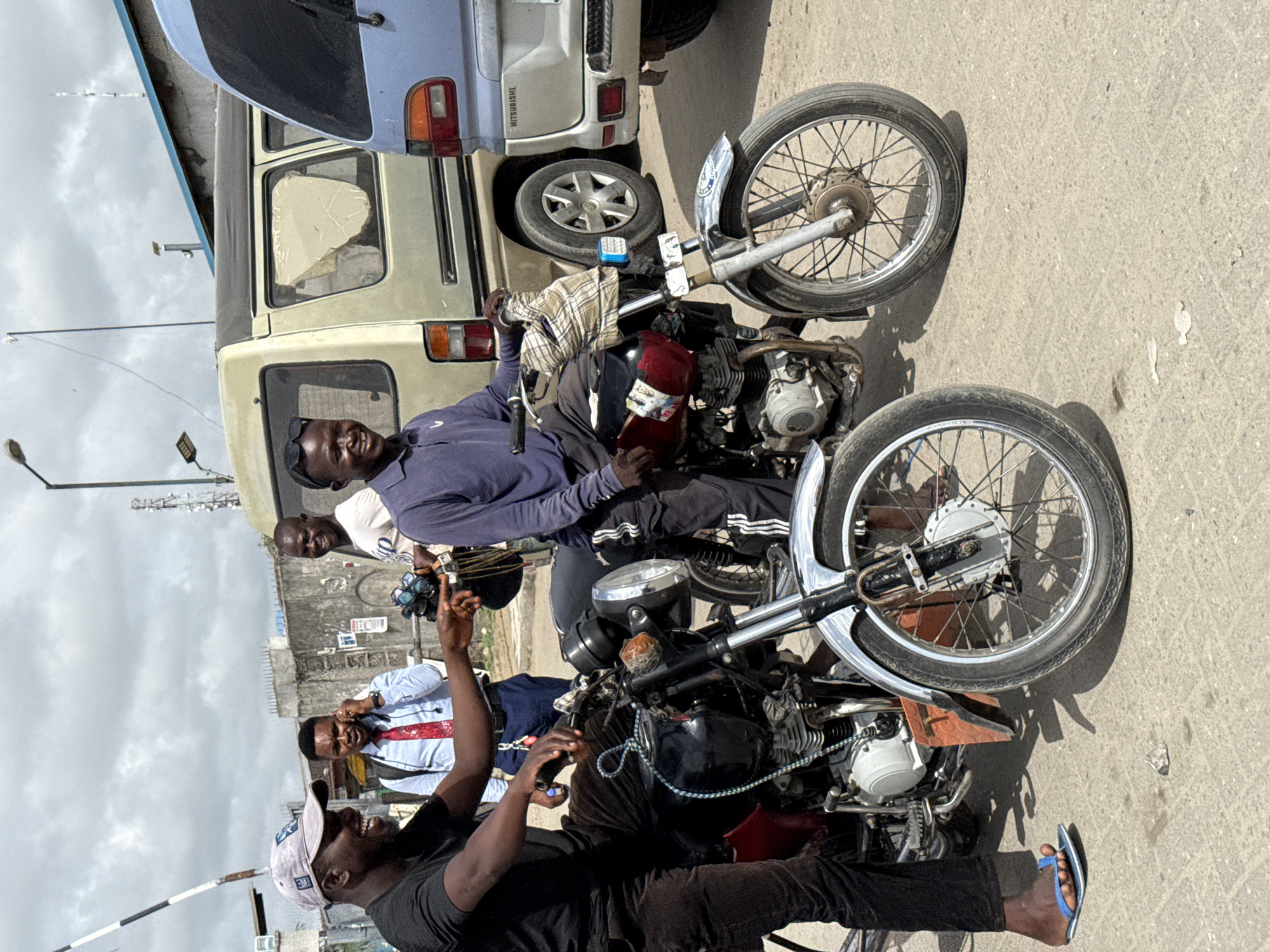 In Lekki, Nigeria, a group of four African men are gathered around two motorcycles parked on a paved road. The motorcycles are facing in opposite directions, with one angled slightly towards the right and the other angled slightly towards the left. A white van is parked in the background, partially obscured by the motorcycle. The men are engaged in conversation, with some gesturing and smiling. The environment consists of urban elements such as buildings, utility poles, and an overcast sky.