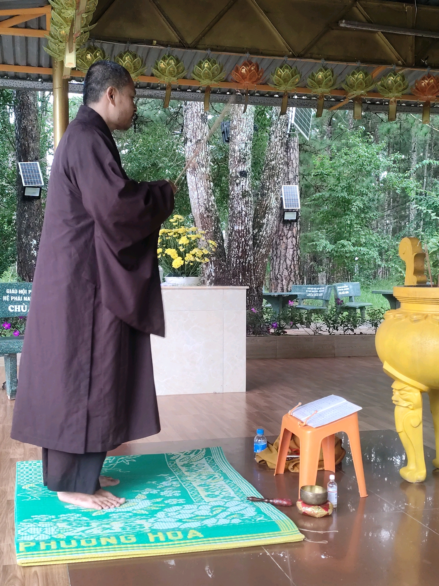 The image depicts a monk standing on a green mat, facing away from the viewer, in what appears to be a Buddhist temple or spiritual site in Da Lat, Vietnam. The monk is wearing a dark brown robe and black trousers. In the background, there are trees, a white structure with a bouquet of yellow flowers, and a yellow decorative urn. Solar panels are visible on some trees, indicating a blend of traditional and modern elements. In the foreground, to the right of the mat, there is an orange plastic stool with a book on it, a small bell, and two bottles of water. The overall setting suggests an outdoor religious practice or ceremony.