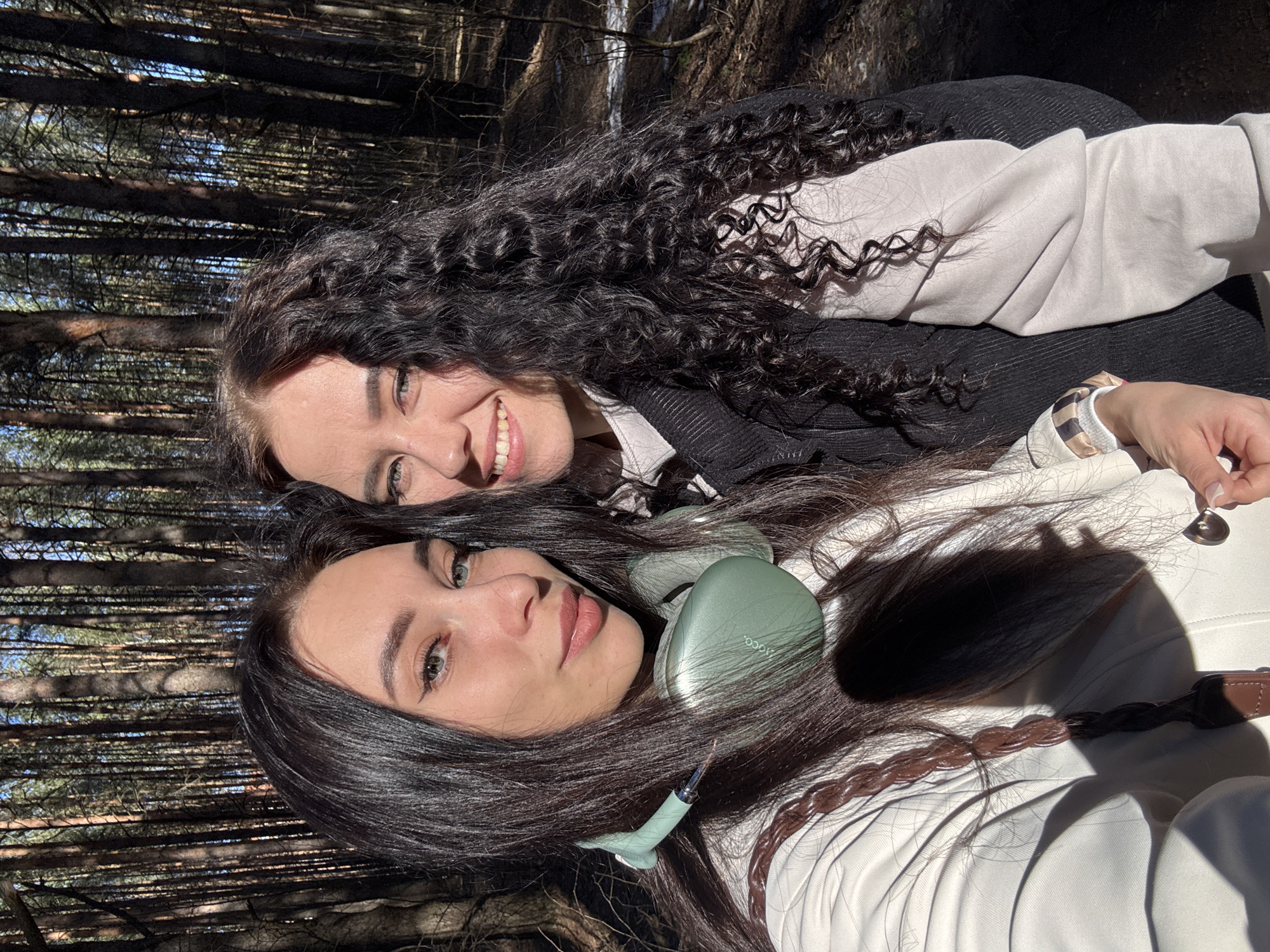 Two young women are posing for a selfie in a forest environment. The woman in the foreground has long, dark hair with a braided accent, light skin, and is wearing light green headphones. The woman in the background has curly, dark hair, fair skin, and is smiling. Both are wearing light-colored jackets. The background shows a dense forest with trees reaching high, filtering sunlight through the canopy. The location is the city of Chaikovsky, Russia.