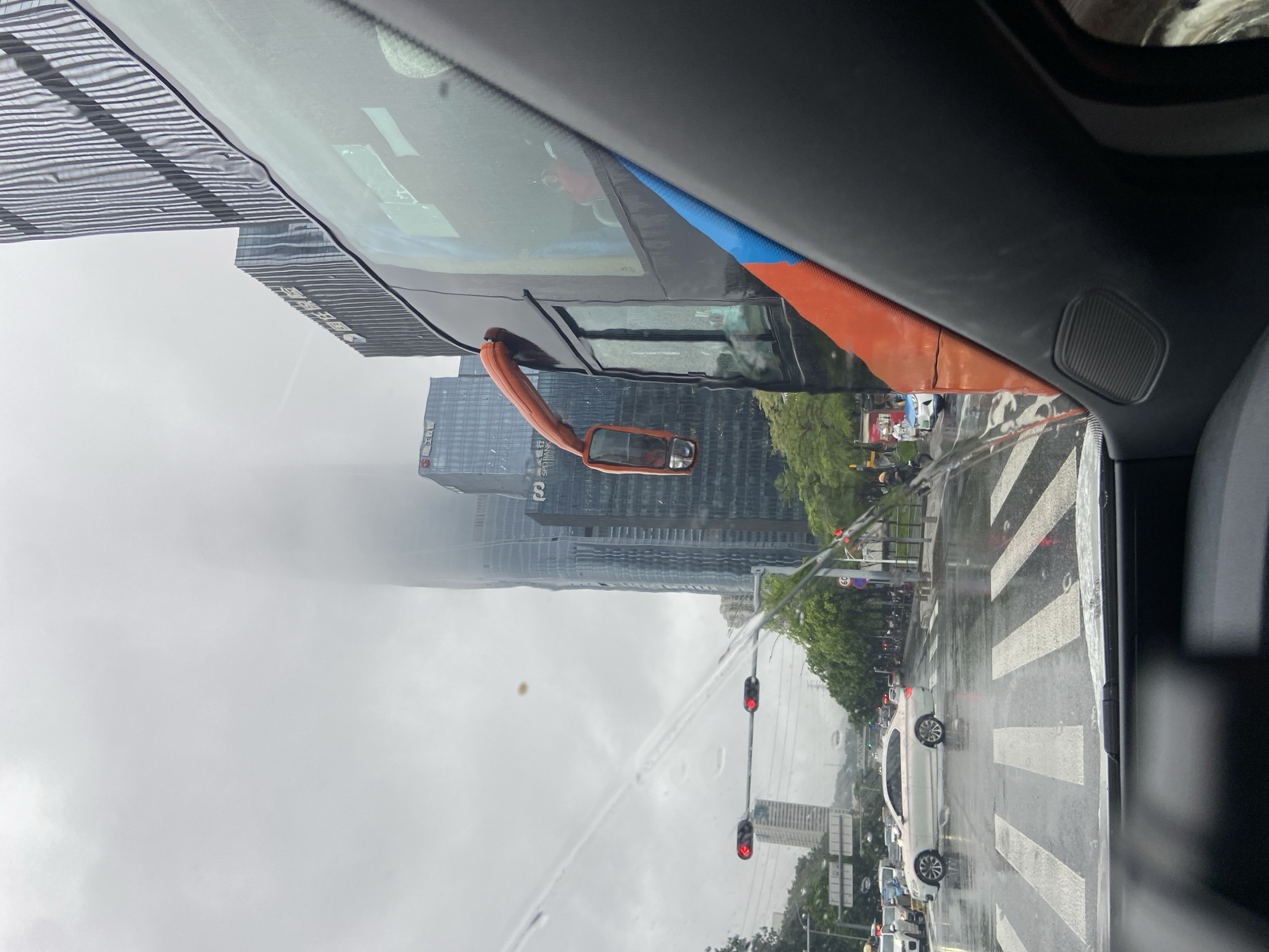 The image depicts a view from inside a vehicle, looking out onto a wet city street in Futian District, China. Raindrops are visible on the windshield, distorting the view of the scene outside. Tall, modern skyscrapers dominate the background, their upper sections obscured by heavy fog or clouds. Traffic lights are red, indicating a stop. A white sedan is positioned on the wet road, in front of a pedestrian crossing. To the right, a portion of a large bus is visible, with an orange side mirror. The sky is overcast and grey. The overall environment suggests a rainy day in a dense urban setting.