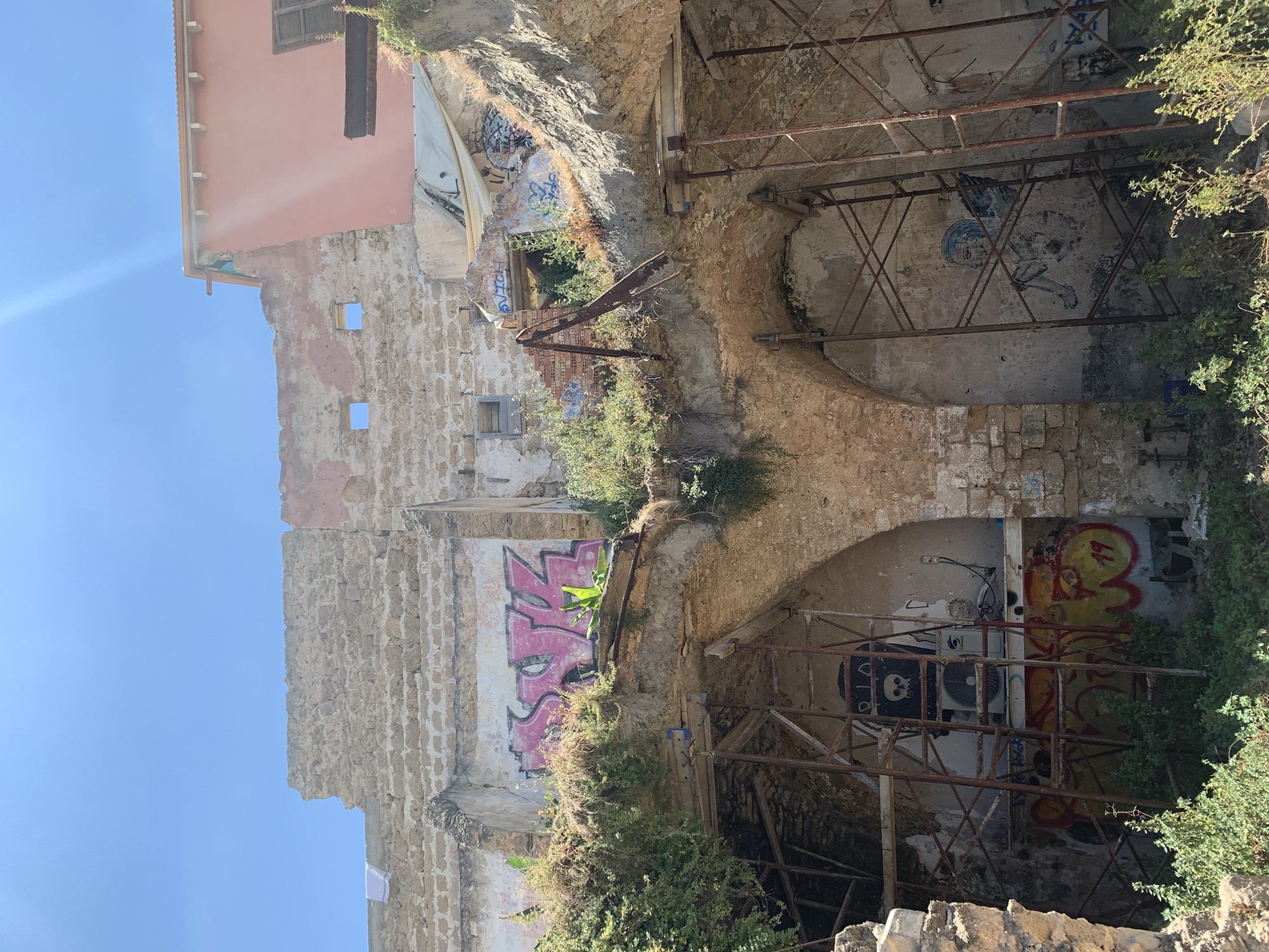 The image shows the partially demolished and graffiti-covered ruins of a building in Tel Aviv, Israel. The structure appears to be made of stone and brick, with some sections still standing and others collapsed. Scaffolding is visible, suggesting ongoing construction or demolition work. Various pieces of graffiti, including tags and drawings, are present on the walls. Lush green vegetation is growing around and within the ruins. The sky is clear and blue.