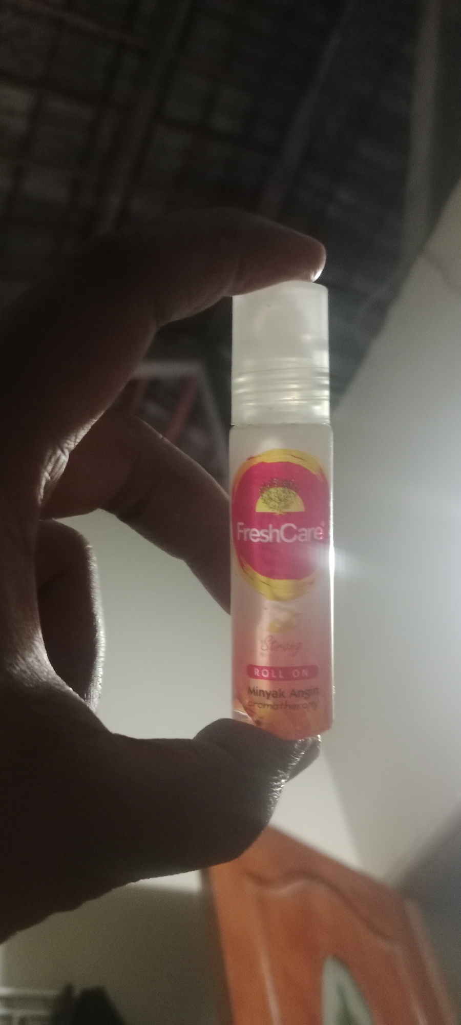 A person's hand holds a small, clear plastic bottle with a white roll-on applicator and cap. The bottle has a pink and yellow label with the text "FreshCare" prominently displayed in white. Below this, the words "ROLL ON" are visible in red, followed by "Minyak Angin" and "aromatherapy" in smaller text. The bottle is positioned in front of a blurry background that appears to be indoors, possibly a room with a ceiling structure and a wall. The lighting suggests an indoor environment, with some shadows cast on the person's hand and the bottle. The image has a slightly grainy quality. The location in Soko, Indonesia, is not discernable from the visual information provided.
