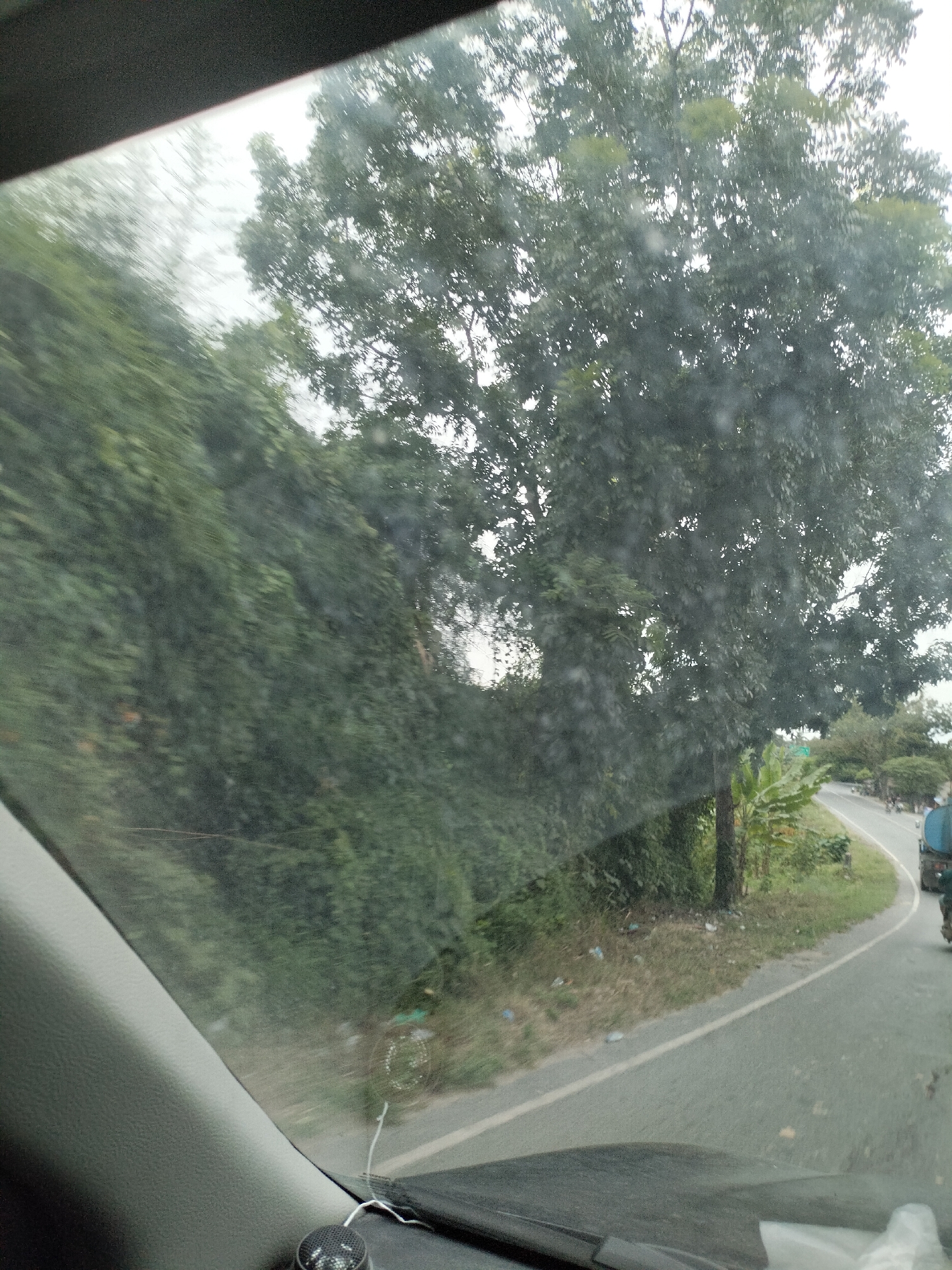 The image, taken from inside a vehicle, shows a view through the windshield. A dense green bush-covered embankment borders the left side of a grey asphalt road that curves to the right. A large tree with dark green foliage stands prominently on the right side of the road, with a thinner tree trunk visible in front of it. Beyond the road, more trees and vegetation are visible, some appearing lush green and others with lighter foliage. A motorcycle is partially visible on the right side of the road, as is a portion of a blue cylindrical object, possibly a water tank on a vehicle. The sky is overcast and appears bright, suggesting a cloudy day. The lower portion of the image shows the dashboard of the vehicle, including a black speaker and a white wire. The image is slightly blurred, indicating movement. The location is identified as Kampung Lama, Indonesia.