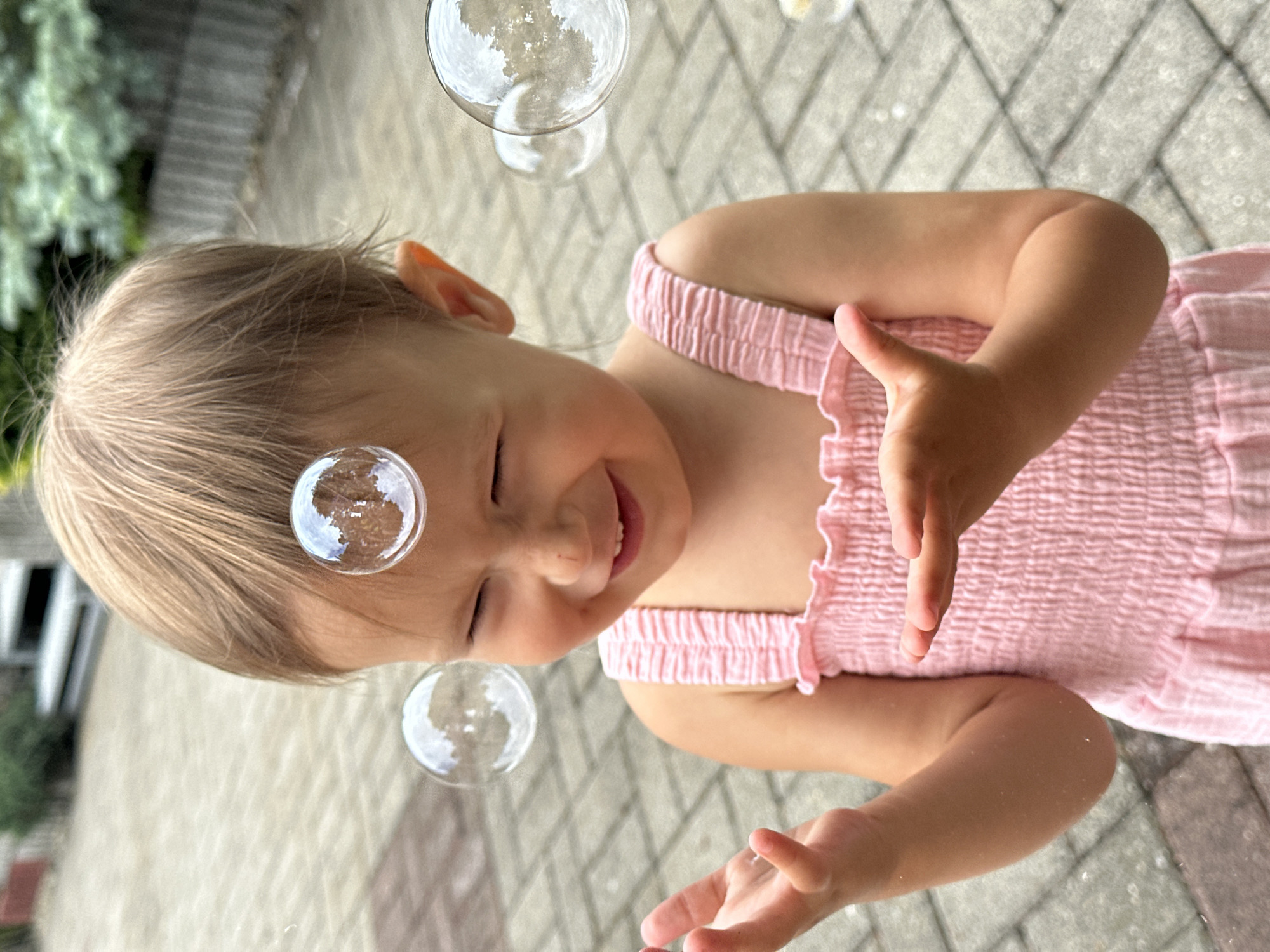 The image depicts a young child with blonde hair, wearing a pink, ruffled dress, outdoors on a paved surface. Several clear, spherical bubbles are floating around the child. The child is smiling with their eyes closed and their mouth open, appearing to be actively engaged with the bubbles. One hand is raised, fingers slightly spread, as if reaching for or interacting with a bubble. Another bubble is close to the child's face, reflecting the surroundings. The background shows a grey, diamond-patterned paved ground and some out-of-focus greenery on the left, suggesting a backyard or garden setting. The lighting suggests it is daytime, likely a sunny day given the reflections in the bubbles. The overall mood is joyful and playful. The provided location context is Wisła, Poland. There is no visible text in the image.