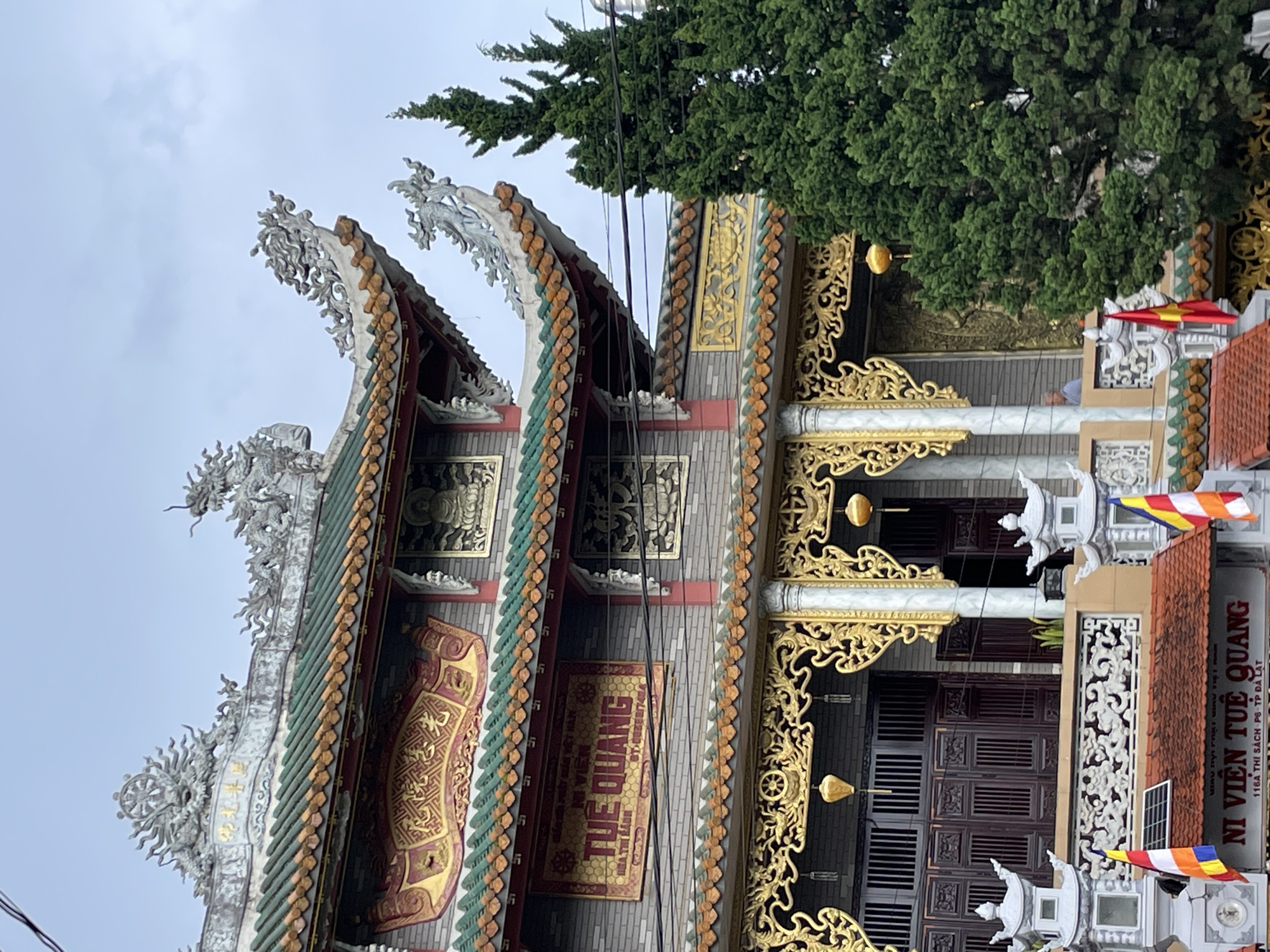 The image shows the facade of a Vietnamese Buddhist temple. The architecture is ornate, with a multi-tiered, curved roof adorned with green and orange tiles. Sculptures of dragons and other mythical creatures decorate the roofline. Intricate golden carvings and reliefs are visible on the building's exterior. A sign with Vietnamese characters is prominently displayed, reading "NI VIÊN TUỆ QUANG". Two Vietnamese flags are positioned in the foreground. A large, dense green tree partially obscures the right side of the temple. The sky is overcast. In the background, a person is visible through a window. The location is identified as Cam Ly - Đà Lạt, Vietnam.