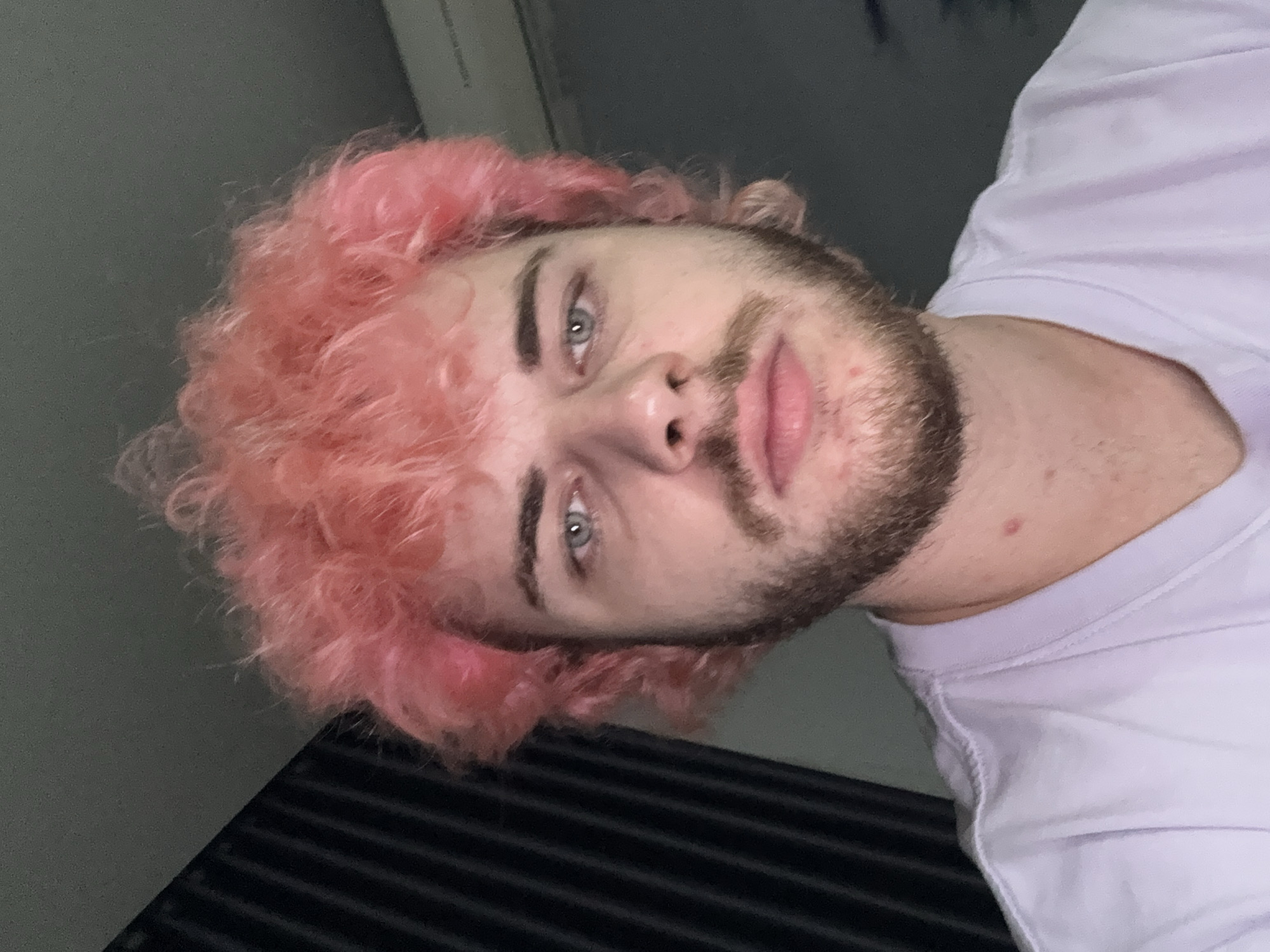 A close-up, tilted portrait captures a young Caucasian male with pink, curly hair and light blue eyes. He has a brown beard and mustache. He is wearing a white t-shirt. The background is a blurry combination of gray and black, suggesting an indoor environment. The presence of Đà Nẵng, Vietnam cannot be confirmed from this image.