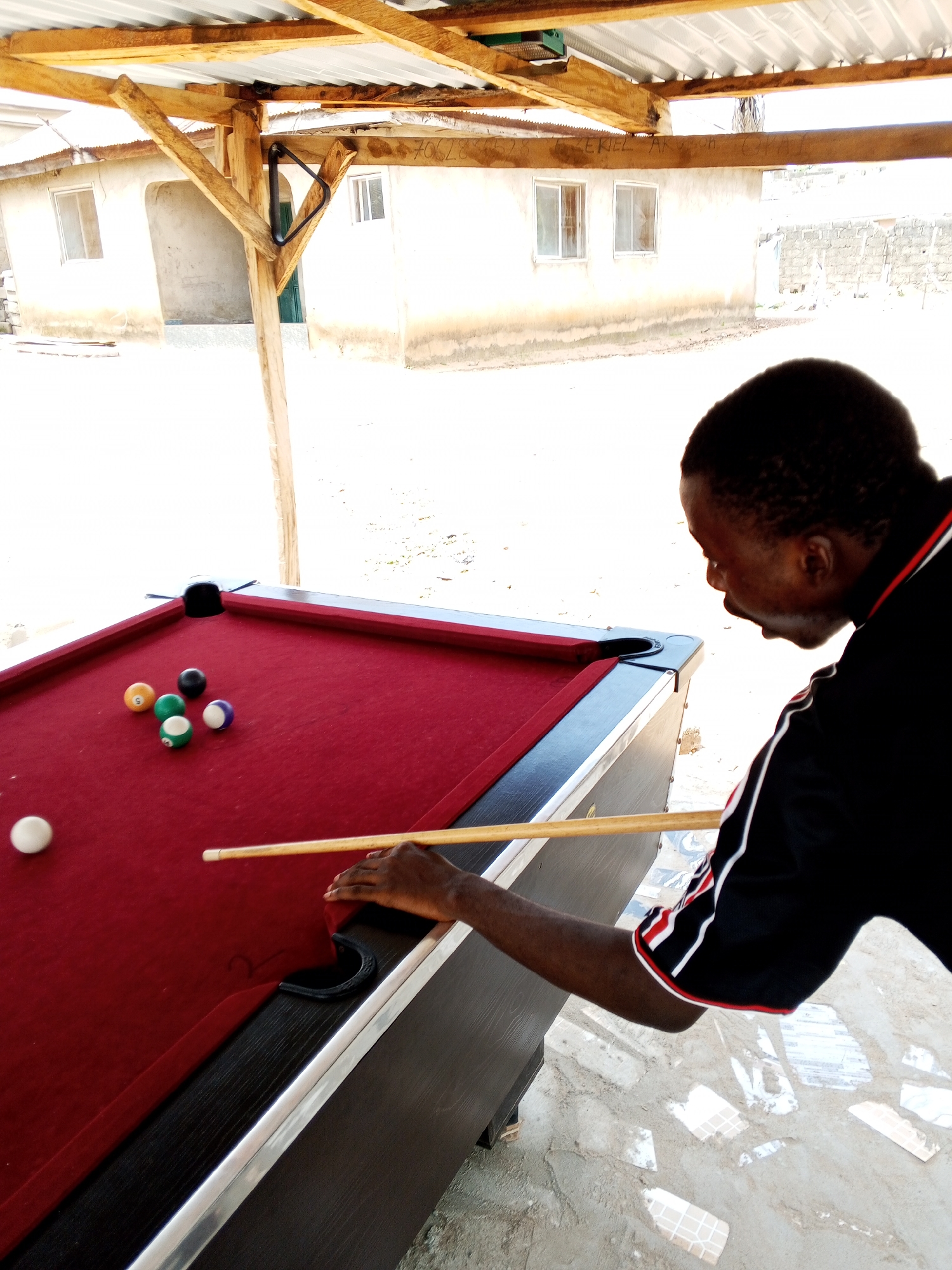 A man is playing pool at a covered outdoor area in Lokoja, Nigeria. The pool table is a standard size with a red felt surface and black pockets. There are several billiard balls on the table, including a white cue ball, a solid orange ball, a solid black ball, a striped green ball with the number 5, a solid green ball, and a striped purple and white ball. The man is dressed in a black shirt with red and white trim. He is holding a pool cue and appears to be in the process of taking a shot. The background shows a building with white walls and windows, as well as some of the wooden support structure for the covered area. The ground is covered in a light-colored material with some scattered debris and patterned tiles. The overall scene suggests a casual setting for playing pool outdoors.