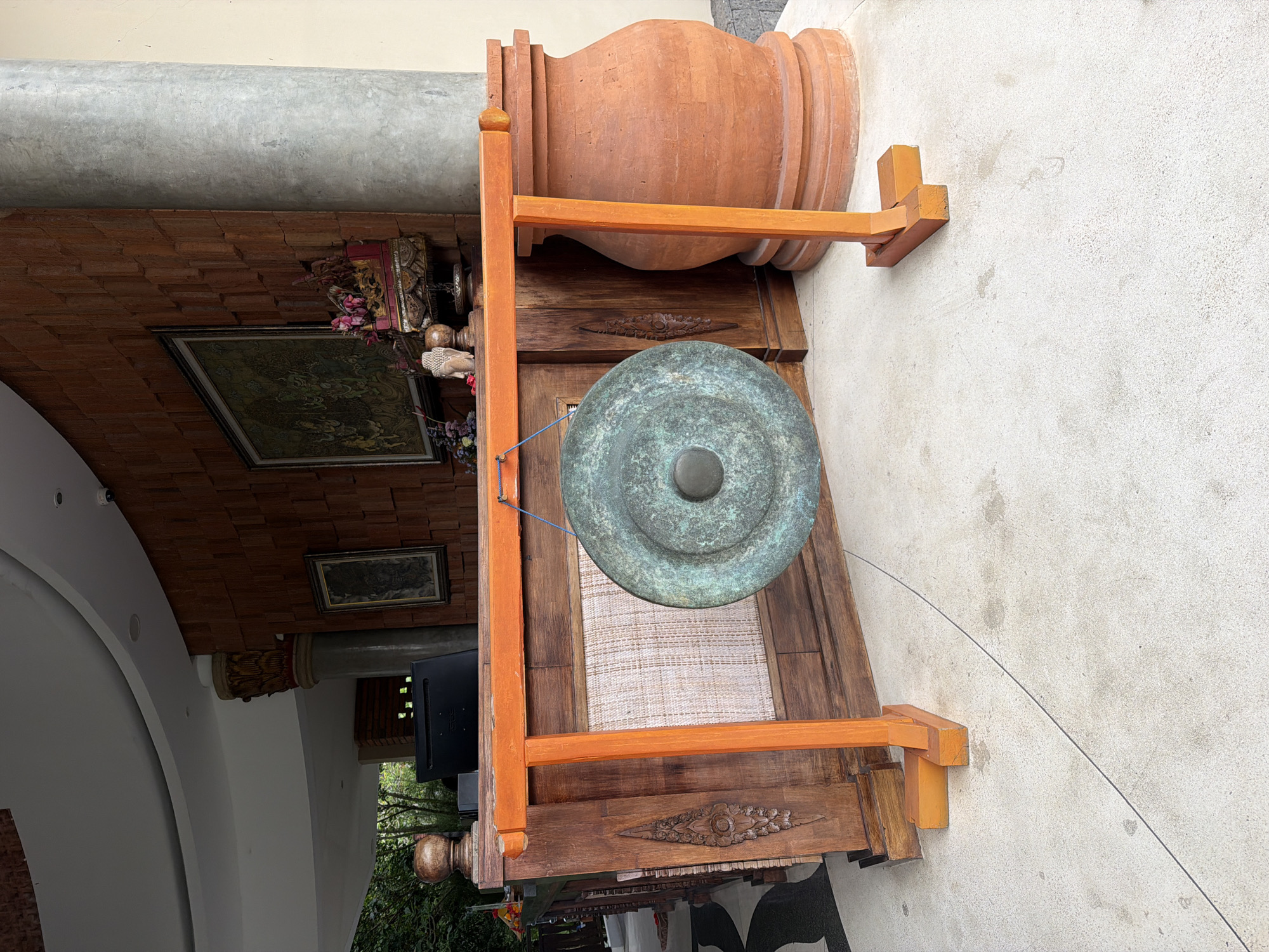 This image shows a gong suspended in a wooden frame, along with a large earthen jar on top of the frame. The setting appears to be in Peliatan, Indonesia, based on the architecture and style of the items. The gong itself is a green-tinted metal disk. The wooden frame is orange and brown, with carved details on the lower front panel. The earthen jar is a reddish-brown color and appears to be resting securely on the top part of the wooden frame.
The setting has brickwork and a framed picture is hanging on the wall. There is a column-like structure next to the brickwork. To the left, there is a section with a dark monitor on a stand, with foliage visible through a window behind it. A white wall is visible on the right.