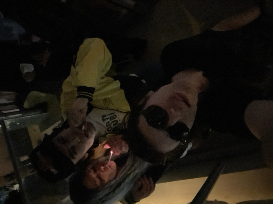 The image depicts three individuals in a dimly lit, close-range setting, identified as an unspecified location within Tel-Aviv, Israel.
In the foreground, a male individual faces the camera, wearing dark sunglasses and a dark t-shirt. He exhibits a slight smile. His left shoulder and arm are partially visible on the left.
Behind him, slightly to the left, a second male is visible. He wears a black baseball cap with a red logo and a yellow and black varsity-style jacket over a white t-shirt featuring a circular logo and partial text. He holds a cigarette-like object to his mouth with his right hand. His left arm is positioned around the shoulders of the female individual beside him.
To the immediate left of the central male, a female individual is present. She possesses long, light-colored hair, styled in a manner consistent with dreadlocks or braids. She is holding a cigarette-like object to her mouth with her left hand while using her right hand to ignite it with a lighter, producing a visible flame. Her gaze is directed towards the ignition point.
The background is dark and lacks discernible details, indicating an indoor or low-light outdoor environment. The individuals are positioned closely, suggesting a social gathering.
