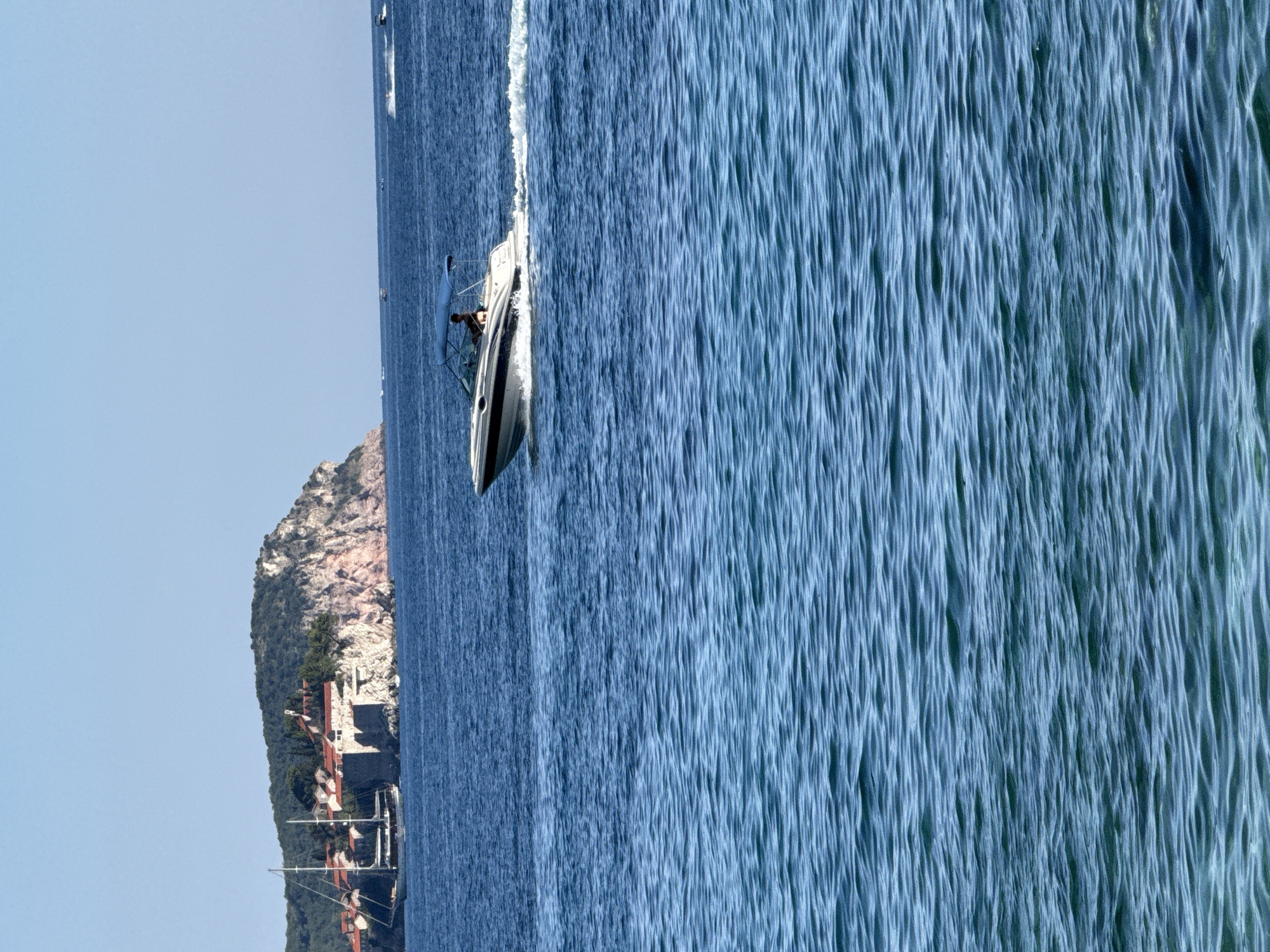 In the city of Rafailovići, Montenegro, a white motorboat with a blue canopy is navigating through the tranquil, blue waters of the Adriatic Sea. The boat is moving from left to right, creating a white wake behind it, and a single person is visible at the helm. In the background, a rocky island with red-roofed buildings and lush green vegetation is visible, with a large sailboat anchored nearby. The sky above is a clear, bright blue, characteristic of a sunny day. The scene captures a serene moment of leisure and travel on the water.