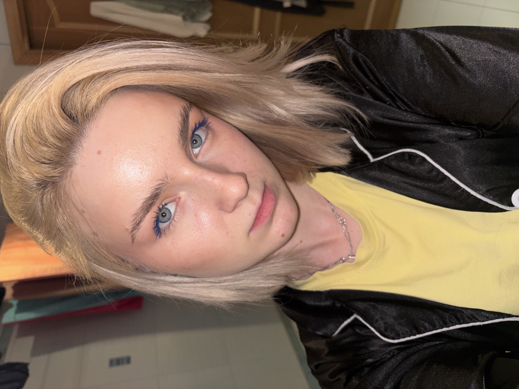 In a close-up, head-and-shoulders selfie, a young Caucasian woman with blonde hair styled in a bob, is positioned centrally. She has light blue eyes and is wearing vibrant blue eyeliner. Her skin appears fair with a subtle sheen, and a few freckles are visible on her cheeks and forehead. She is wearing a yellow t-shirt and a black satin jacket with white piping. A silver chain with a pendant rests on her neck. The background is blurred, showing what appears to be laundry hanging on a rack to the left, and a wooden door and more hanging laundry to the right. The lighting suggests an indoor environment.