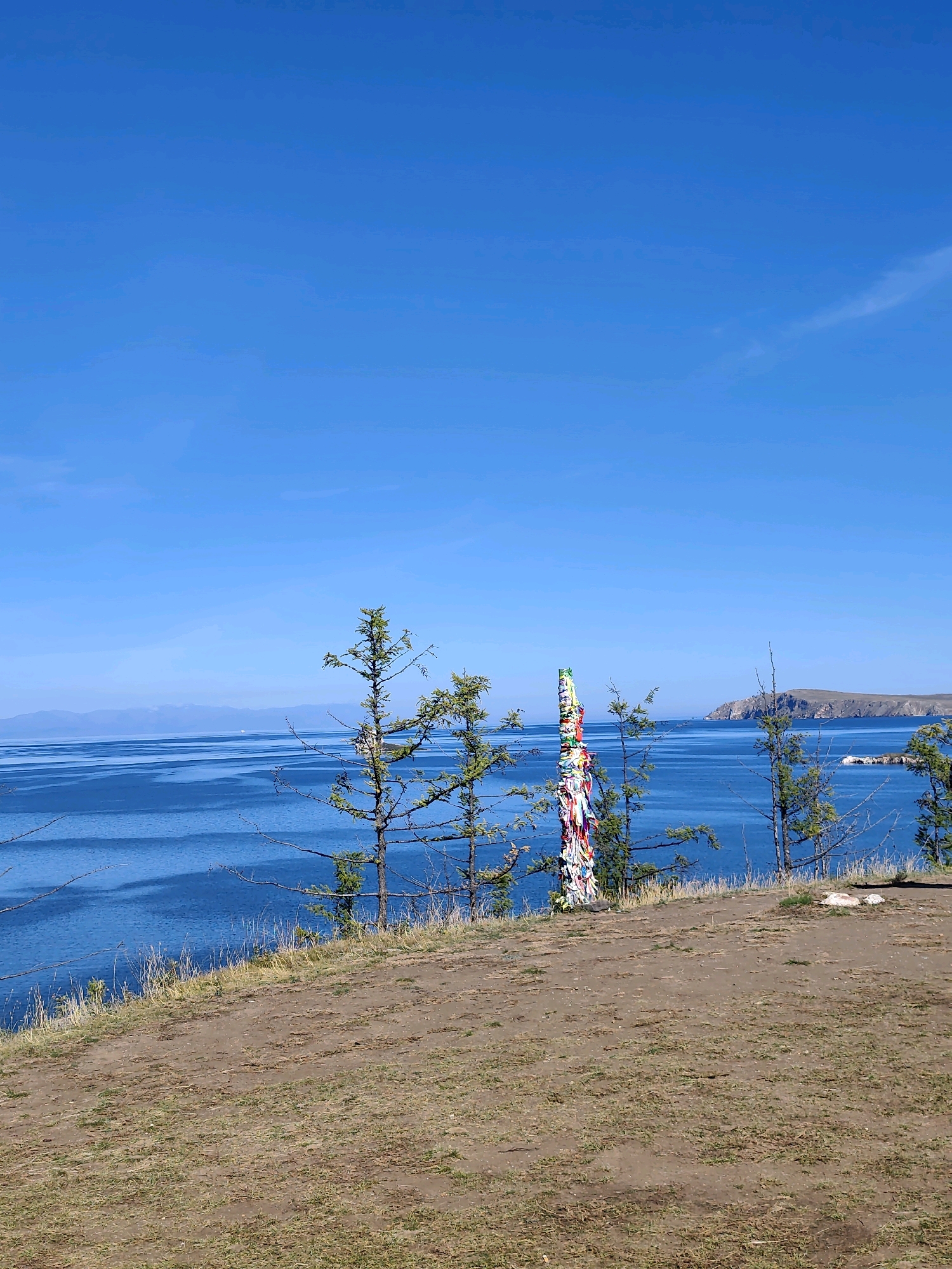 This is a landscape photograph taken in Kharantsy, Russia. The image captures a clear blue sky above a vast expanse of bright blue water. The water is calm, with gentle ripples reflecting the sunlight. In the foreground, there is a dirt path with sparse, dry grass growing on it. Several tall, slender evergreen trees are scattered along the edge of the path, their branches reaching towards the sky. Prominently positioned in the middle ground is a tall, slender wooden pole adorned with numerous colorful ribbons tied around it. These ribbons are of various colors, including white, red, blue, and yellow, and they flutter gently in the breeze. Beyond the water, a distant, hazy mountain range is visible, meeting the horizon under the bright sky. On the right side of the water, a rocky island or peninsula juts out into the water, its slopes covered with vegetation. The overall impression is one of a serene and natural environment, likely a scenic overlook.