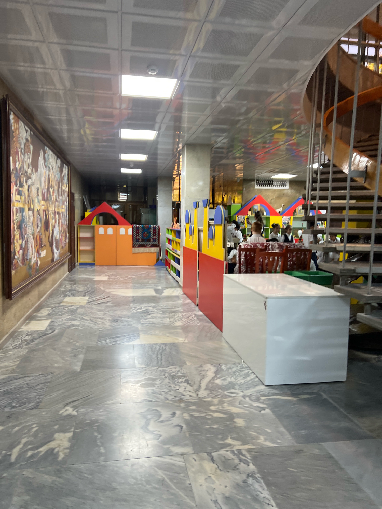 An interior shot captures a hallway with a marble floor. To the left, a large framed painting hangs on the wall. Further down the hallway, a colorful play structure resembling a house is set up. On the right, a spiral staircase with wooden steps and metal railings ascends. Several people, some appearing to be children in uniforms, are visible near the staircase and behind colorful shelving units. The ceiling features square panels with recessed lighting.