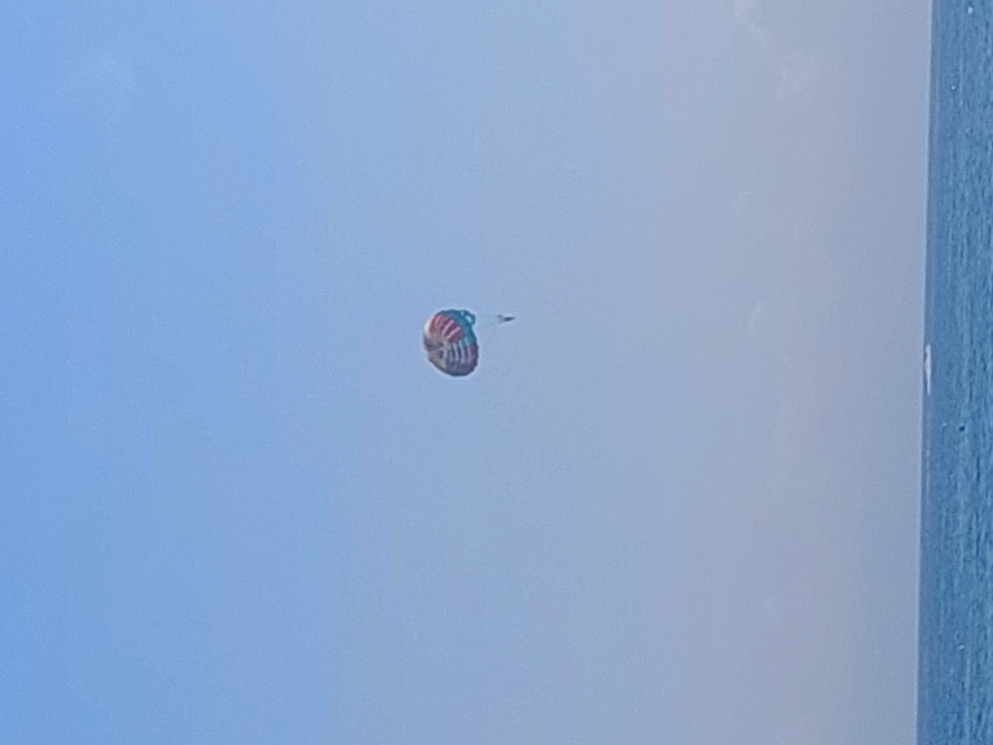 A single parasailer is visible high in the sky, against a clear, pale blue sky. The parasail is multi-colored with red, white, and blue sections. The person being towed is a small, dark silhouette hanging beneath the parasail. Below the sky, the deep blue ocean stretches across the bottom of the frame, with a distinct horizon line. The water appears calm with small ripples. There is no visible indication of the city of Sozopol, Bulgaria, in this image.
