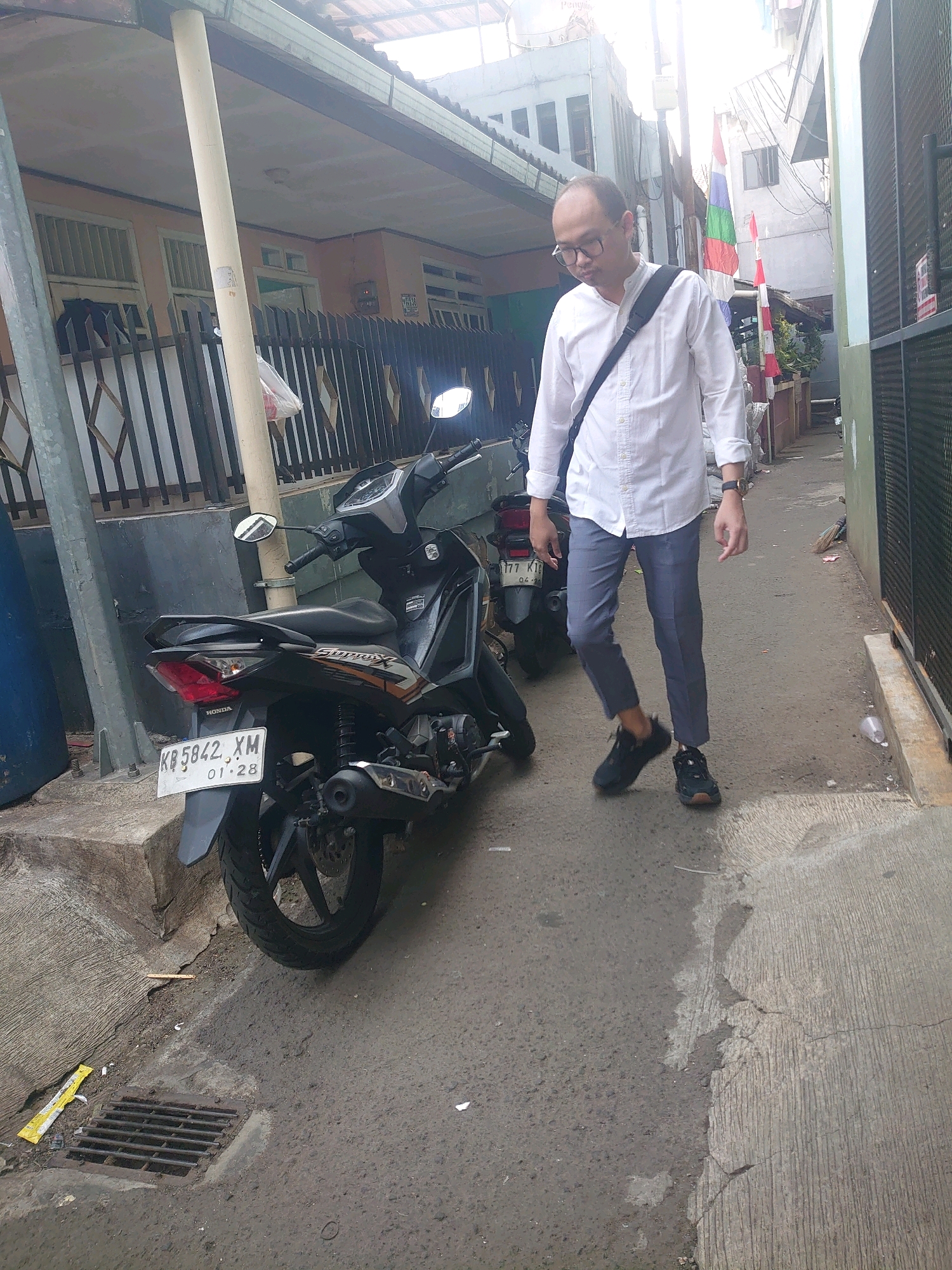 A man wearing a white button-down shirt, grey pants, and black sneakers walks down a narrow street. He has a black messenger bag slung across his chest. To his left, a black motorcycle is parked on the side of the street. The street is paved with asphalt and concrete, with a metal grate covering a drainage ditch. Buildings line the street, with some featuring a light pink facade. Indonesian flags are visible hanging from a building in the background. The scene appears to be in an urban residential area.