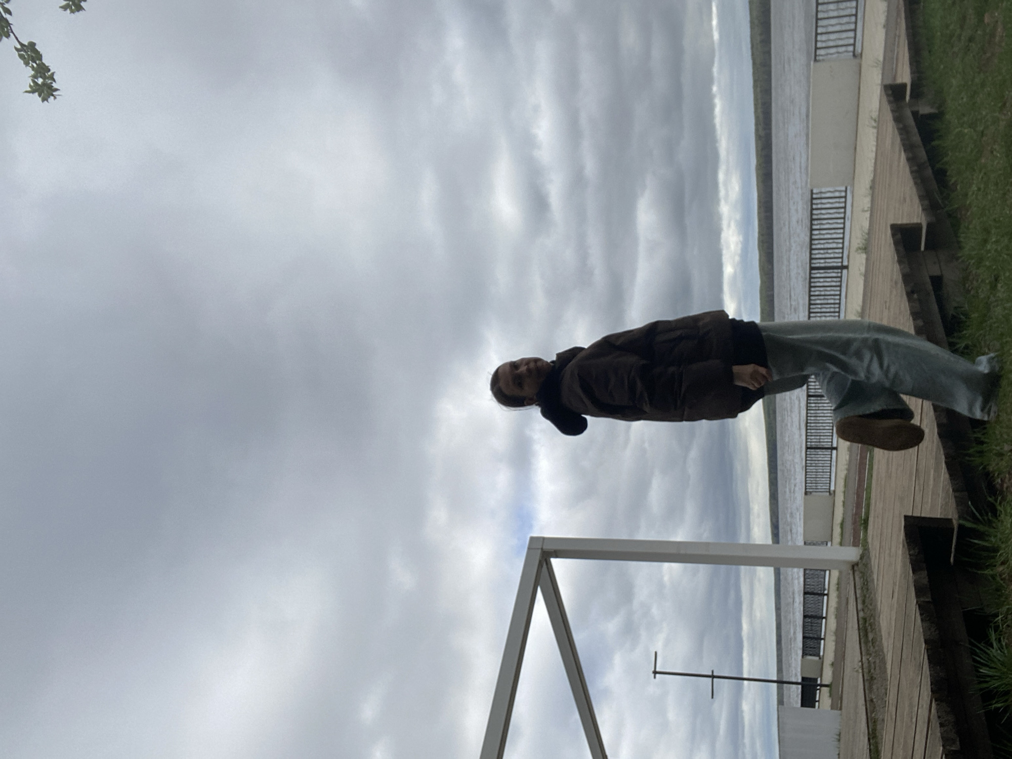 A person stands on a wooden boardwalk next to a body of water on a cloudy day. The boardwalk is next to a grassy area. In the background is what appears to be an urban area. A white metal architectural structure is to the left.
Although the location can be surmised based on the prompt, there is nothing in the image itself that specifically identifies it as being in Chaikovsky, Russia.