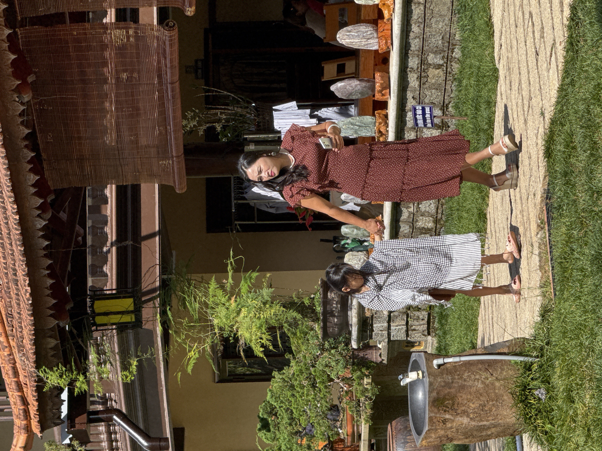 A woman in a brown polka dot dress and a young girl in a black and white checkered sarong walk side-by-side on a stone path in a garden setting in Da Lat, Vietnam. The woman holds her phone in her right hand and is looking to her left. The girl, holding onto her sarong with both hands, is looking down. The garden features a stone sink with a faucet on the left, potted bonsai trees in the background, and decorative stones on display. The architecture suggests a Vietnamese style with a tiled roof and wooden accents. The lighting indicates it is daytime, likely sunny, given the shadows.