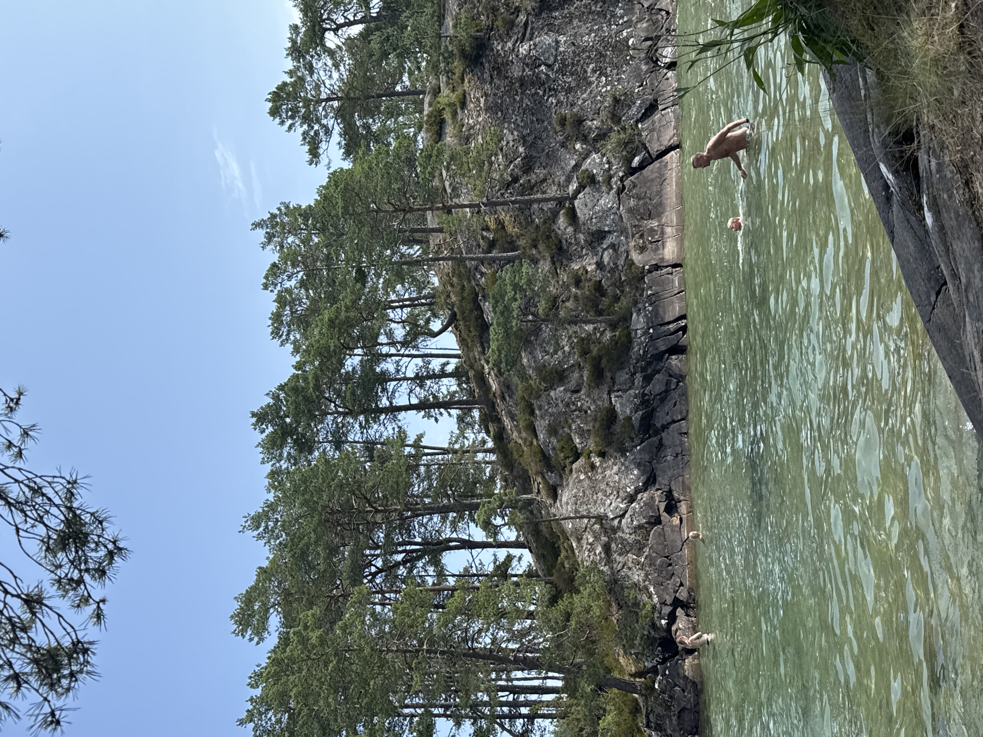 The image depicts a scenic natural environment with a body of water and a rocky, tree-covered hillside. In the water, there are at least three individuals. One person, appearing to be an adult male, is standing in the water and looking towards the right of the frame. Another individual, also appearing to be an adult, is swimming further back in the water. A third person is partially visible in the water near the left edge of the frame. The water is a greenish hue and appears somewhat shallow near the edge. The background features a steep, rocky incline densely populated with tall, slender pine trees. The sky above is clear and blue. The scene suggests a recreational activity, likely swimming or enjoying a natural landscape. This location is likely within or near Karlsborg, Sweden, given the natural features and clear sky typical of the region during warmer months.